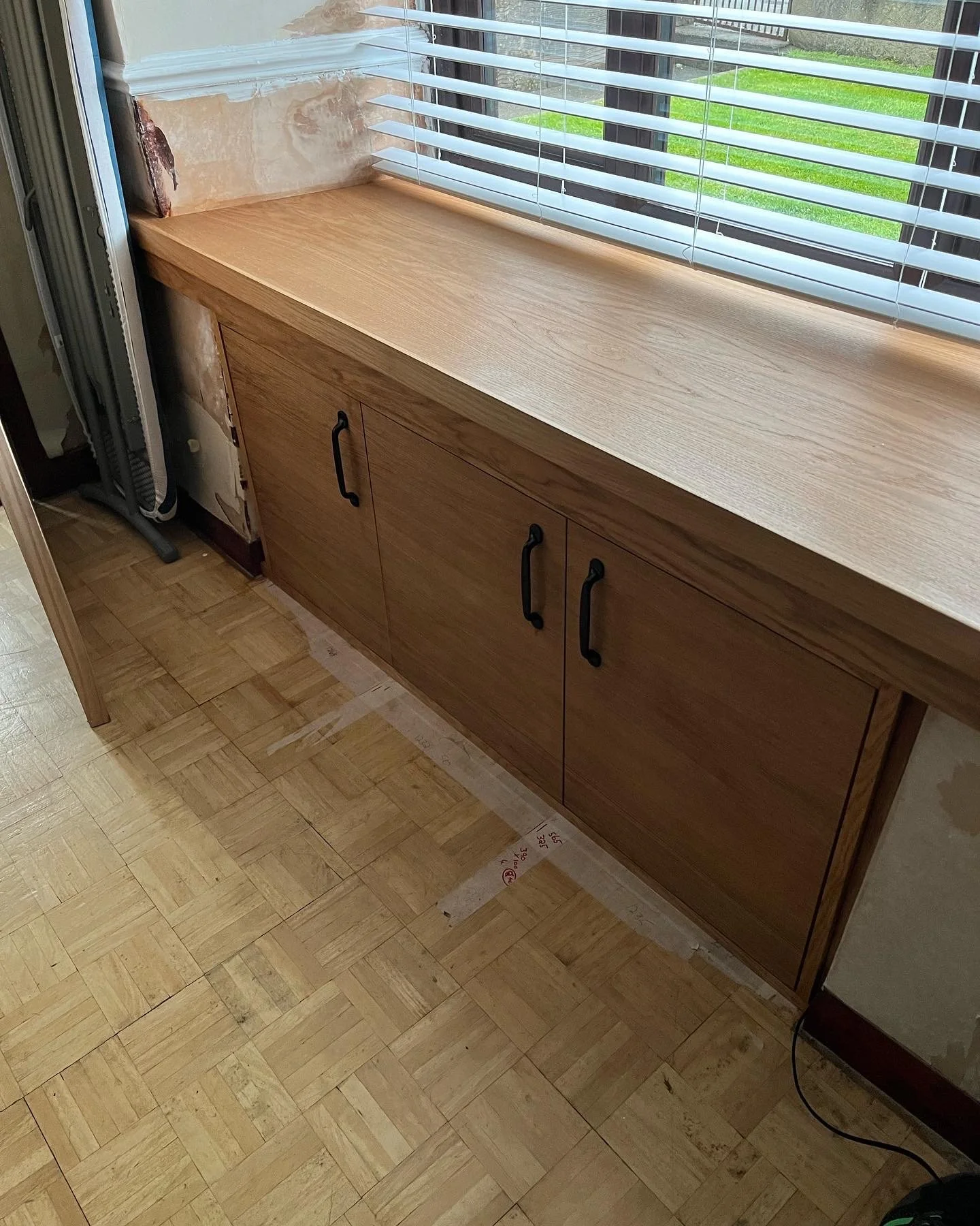 A wooden cabinet with black handles beneath a window with white horizontal blinds, in a room with light-colored parquet flooring and a partially exposed wall section.