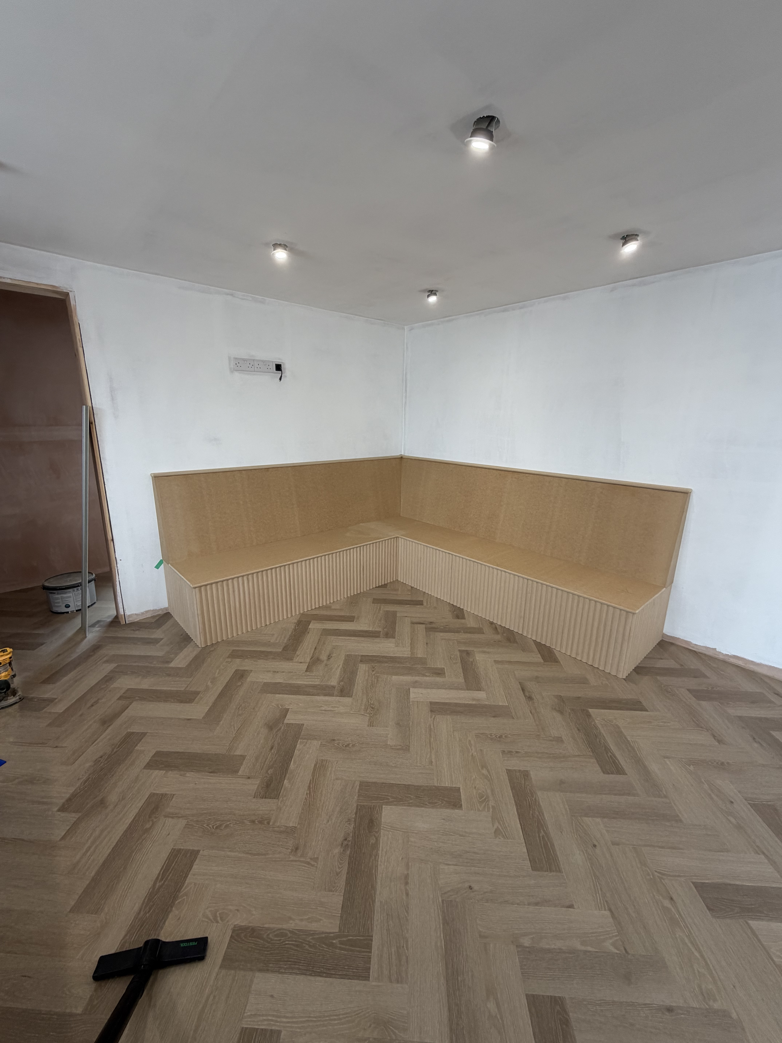 A room with a built-in corner wooden bench with vertical slats on the lower part, white walls, a partly visible door frame, ceiling recessed lights, and a wooden herringbone-patterned floor. A cordless vacuum is on the floor in the foreground.
