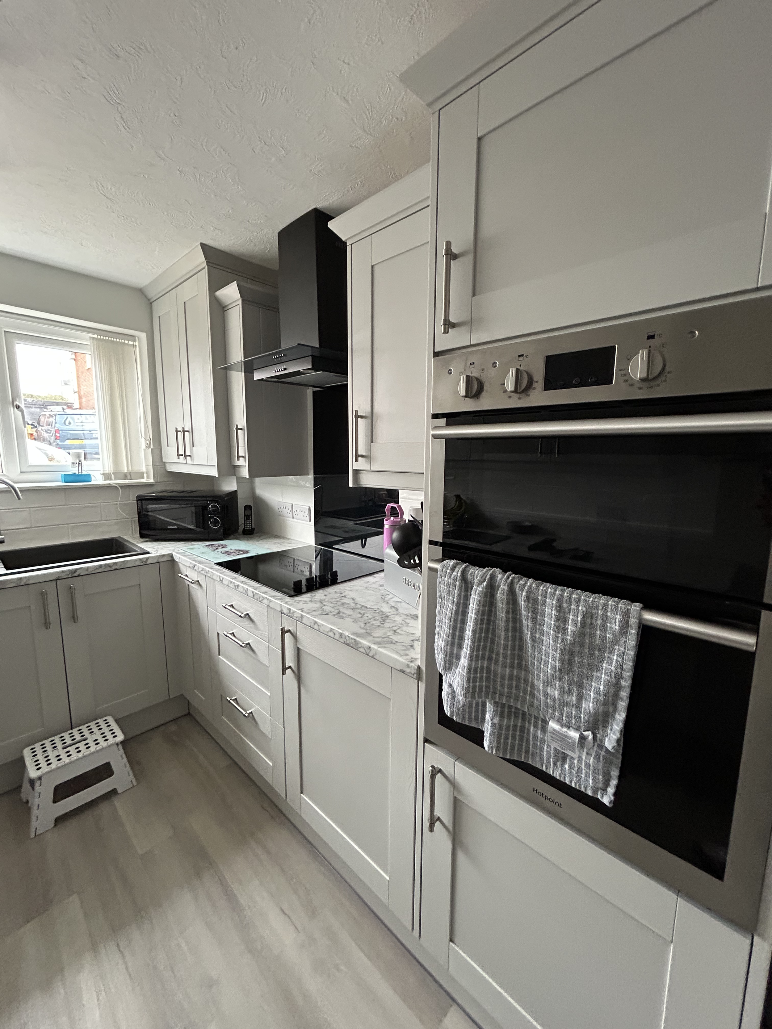 A modern kitchen with white cabinets, a marble countertop, a black oven and microwave, a window with a view outside, a step stool, and various kitchen items including a pink water bottle and a dish towel hanging on an oven handle.