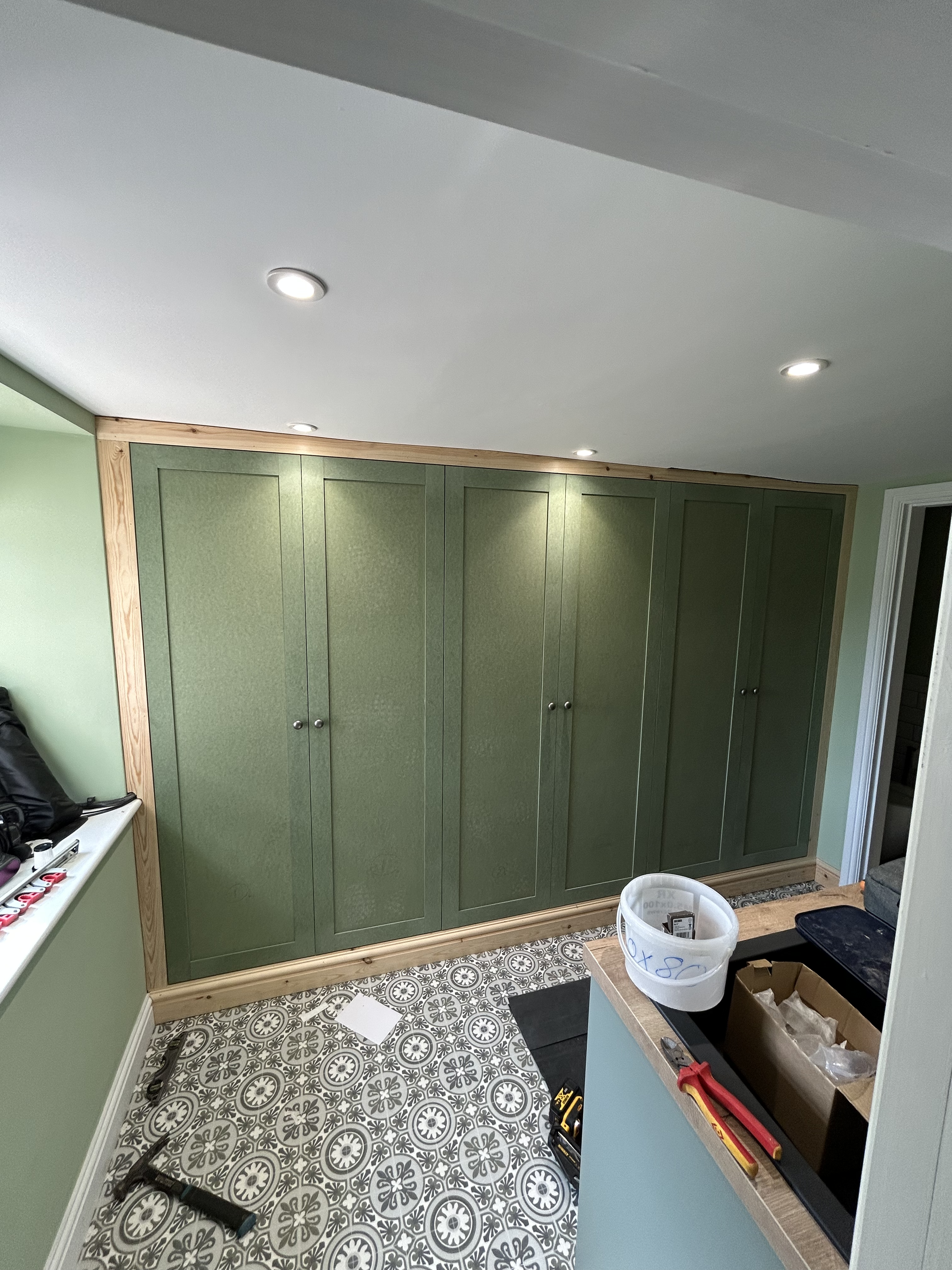 A room with a large green wardrobe with multiple doors, a patterned tile floor, and a ceiling with recessed lights. There is a white countertop with tools and supplies, including a toolbox, a plastic bucket, and a cardboard box. The room appears to b