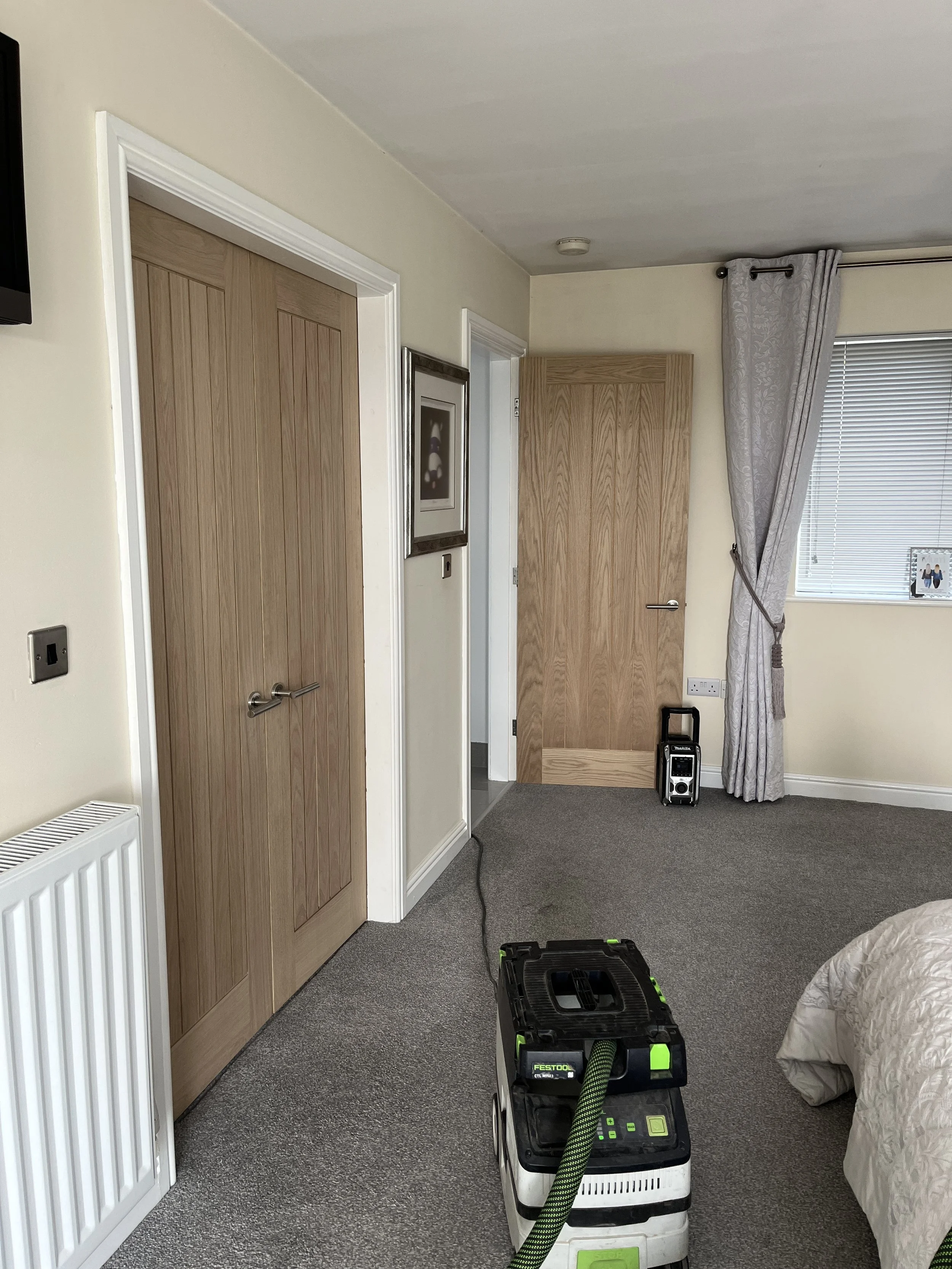 A room with beige walls, a closed wooden door, a window with blinds and a curtain, a picture frame on the wall, a black and green portable vacuum cleaner on the carpet, and a partially visible bed with a cream-colored blanket.