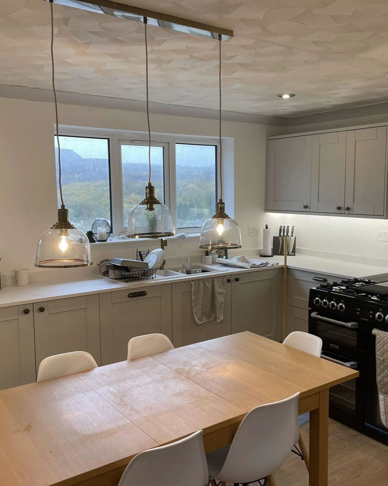 A modern kitchen with white cabinets, a window with a view, and three hanging pendant lights over the countertop. There is a stove, a dish rack with dishes, and various kitchen utensils. In the foreground, there is a wooden dining table with white ch