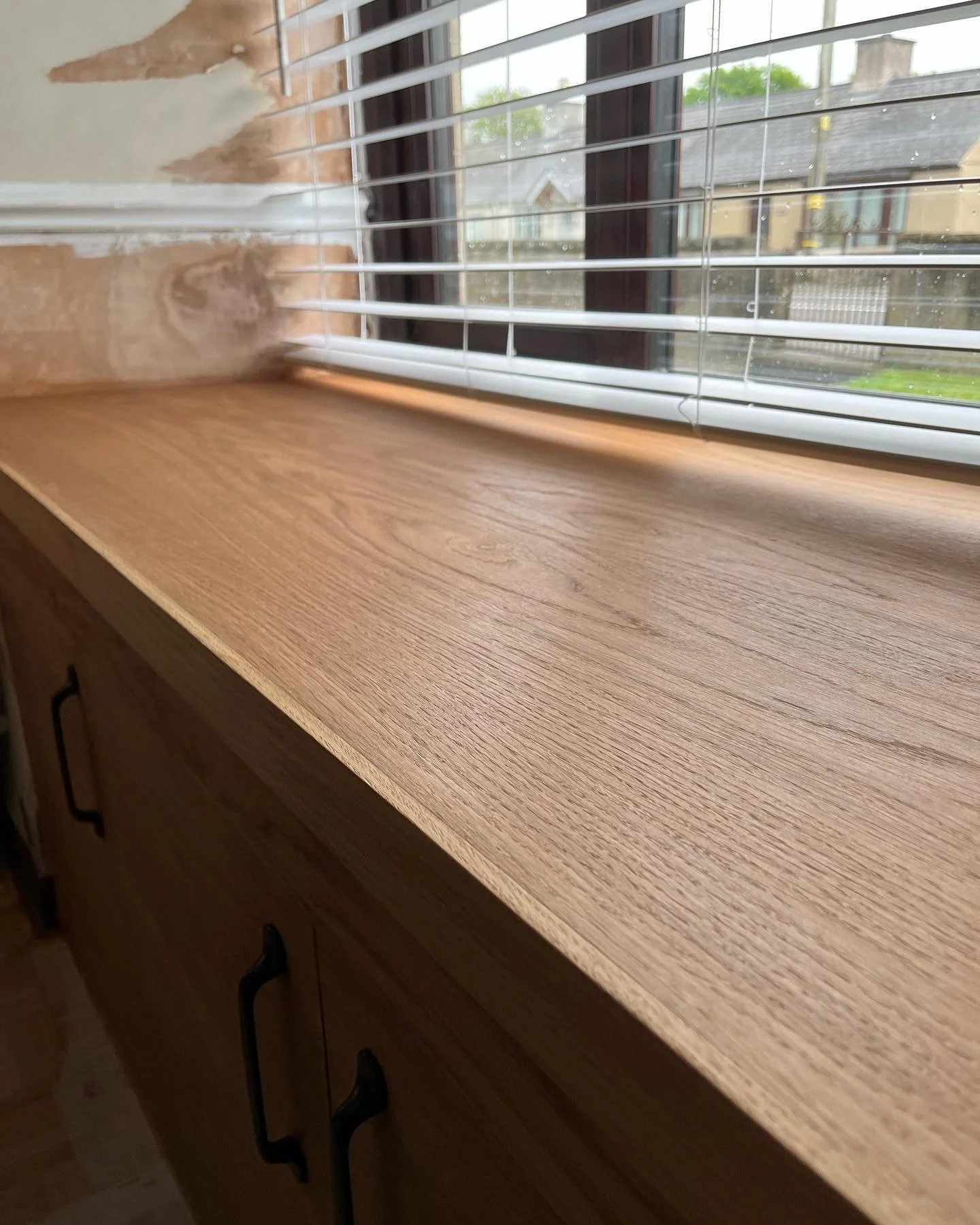 A wooden windowsill next to a window with white blinds, with peeling paint on the wall and a view of houses and trees outside on a rainy day.