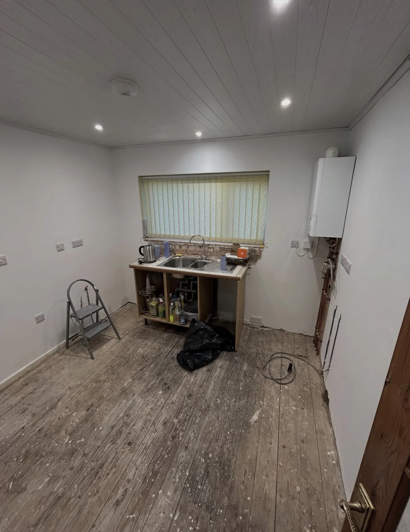 A nearly empty kitchen with white walls and a wooden floor. There is a small counter with a sink, some supplies underneath, and a window with a yellow patterned curtain. There is a folding chair, some tools, and a black trash bag on the floor. A boil