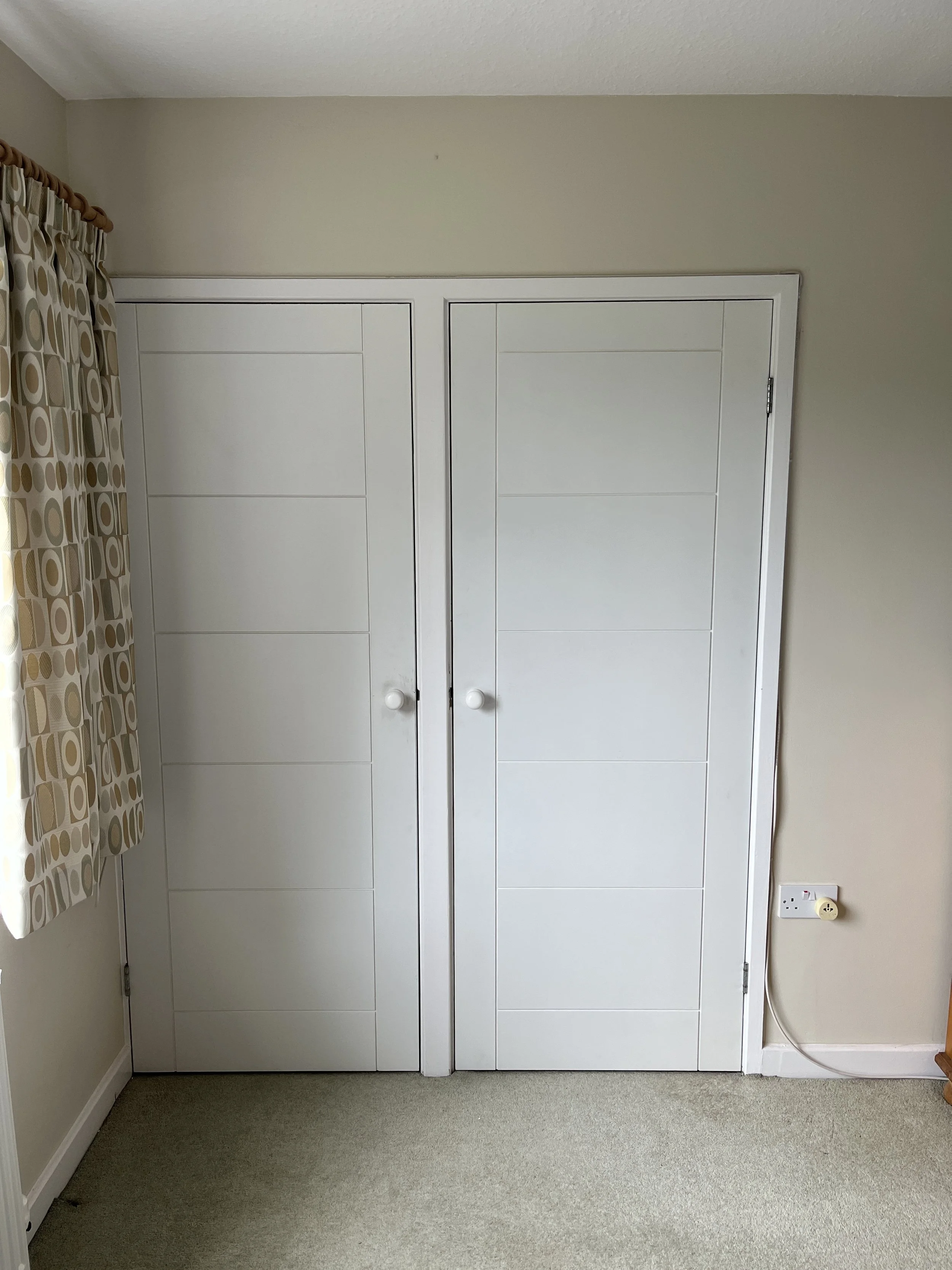 White double wardrobe with two doors and round handles, against a beige wall. A power outlet and plug are visible on the right side near the floor, with a beige carpet on the floor and a window with patterned curtains to the left.