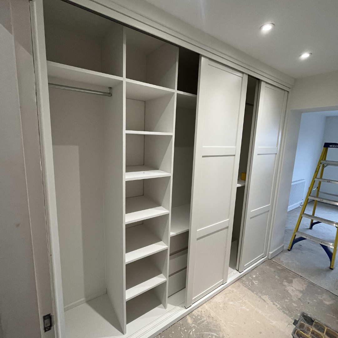 White built-in closet with sliding doors, open to show shelving and hanging rod, in a room with recessed ceiling lights and a yellow ladder in the background.