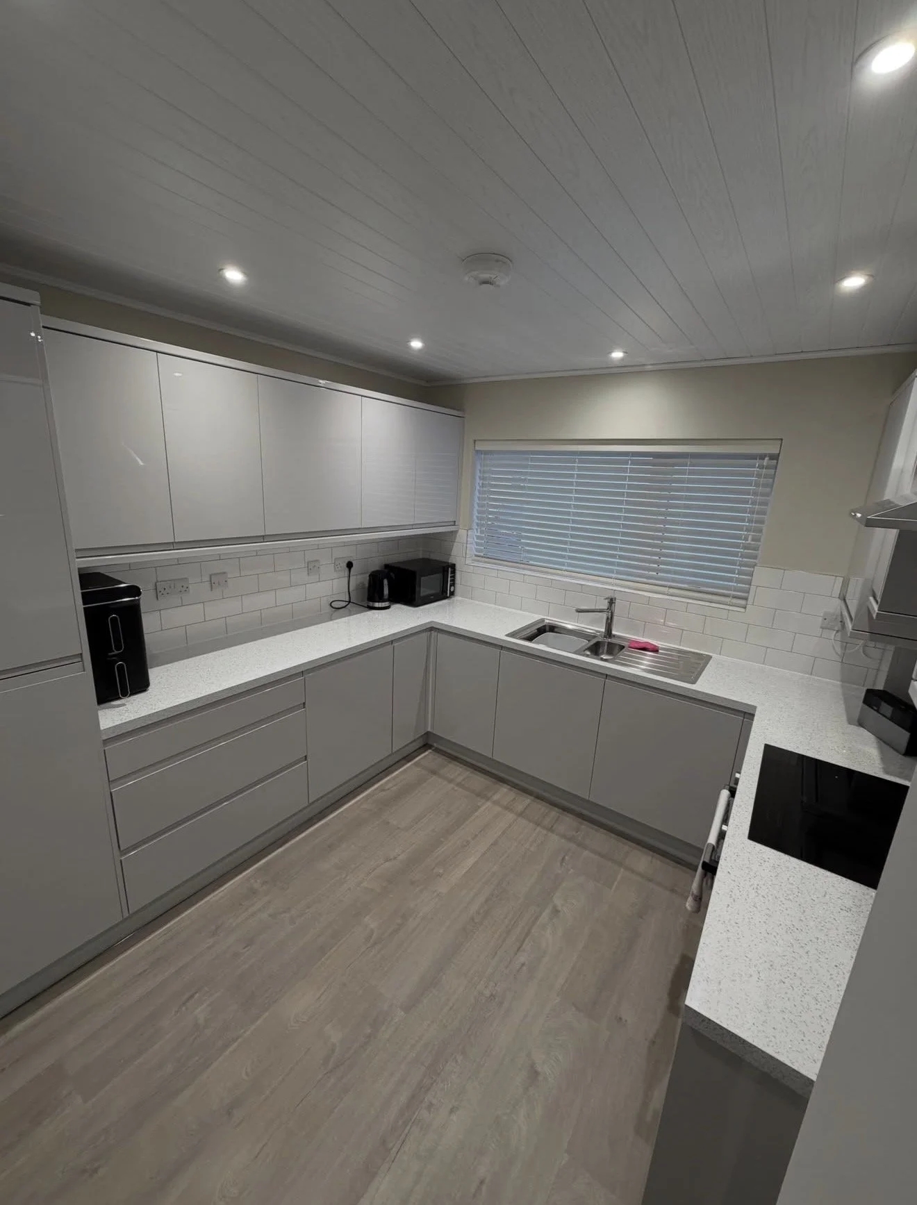 Modern kitchen with white cabinets, grey countertops, a stainless steel sink, and a window with blinds. Kitchen appliances include a microwave, a kettle, and a toaster. The ceiling has recessed lighting.