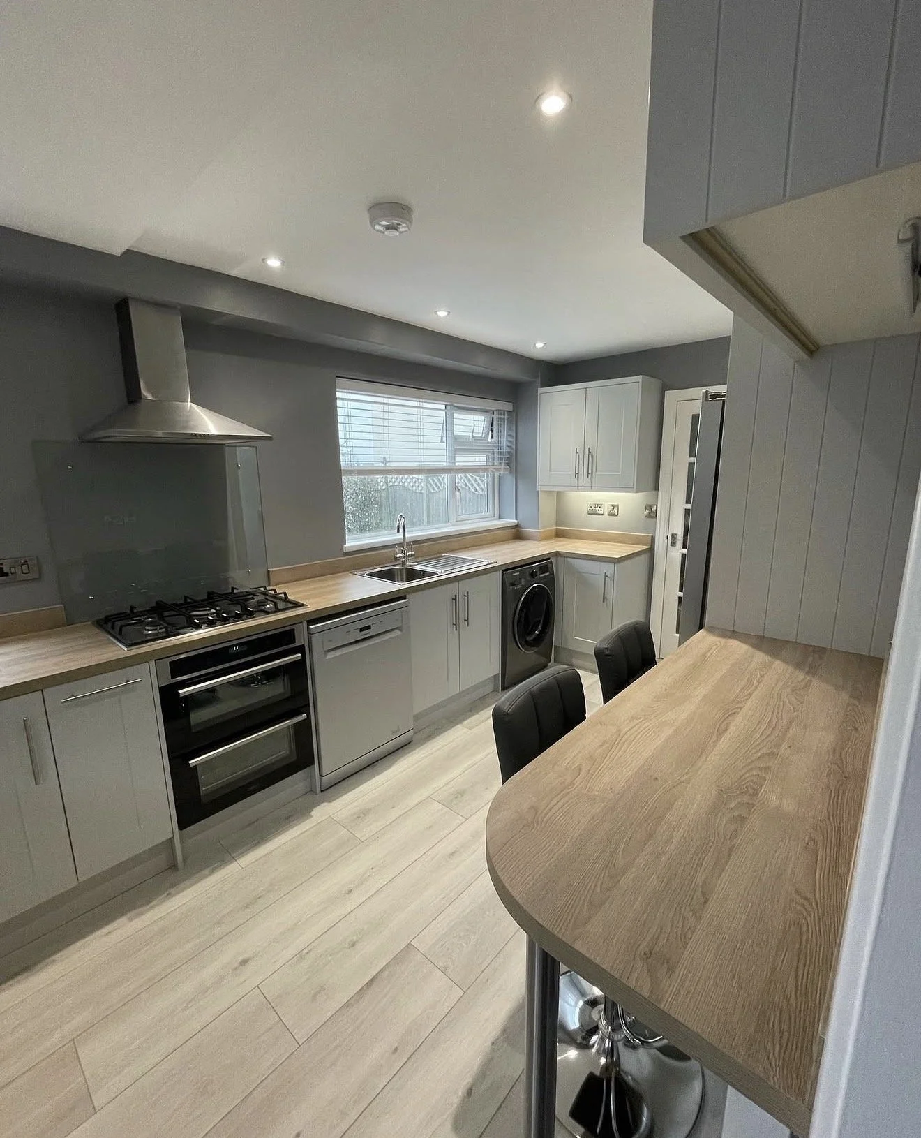 Modern kitchen with gray cabinets, wooden countertops, a gas stove, oven, dishwasher, washing machine, and a window above the sink, with seating at a wooden bar counter.