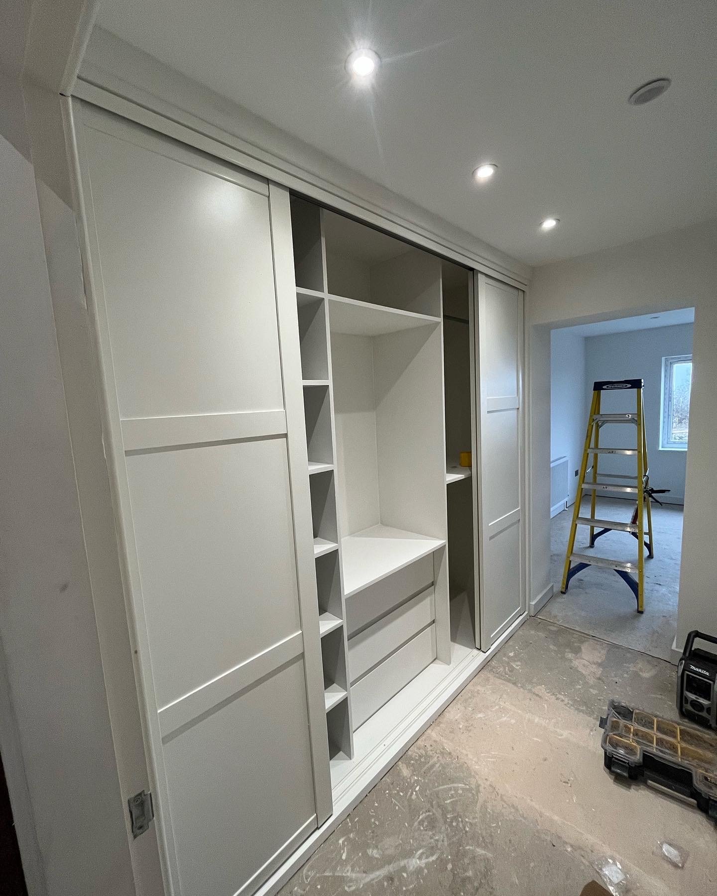 White built-in shelves and cabinets under construction in a room, with a yellow step ladder nearby and construction tools on the floor.