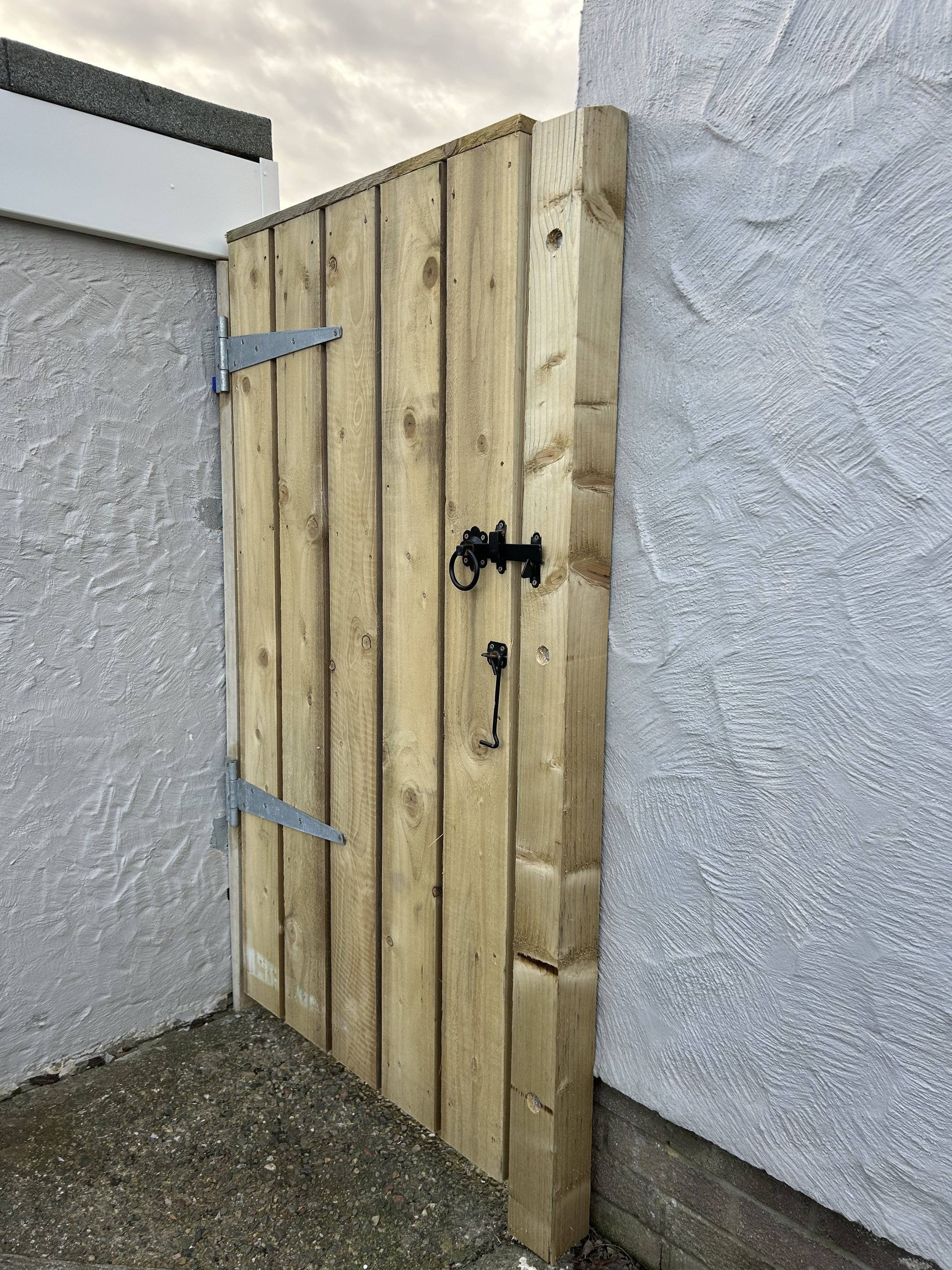 A newly installed wooden gate with black hardware, attached to a textured white wall on a gravel surface, under a cloudy sky.