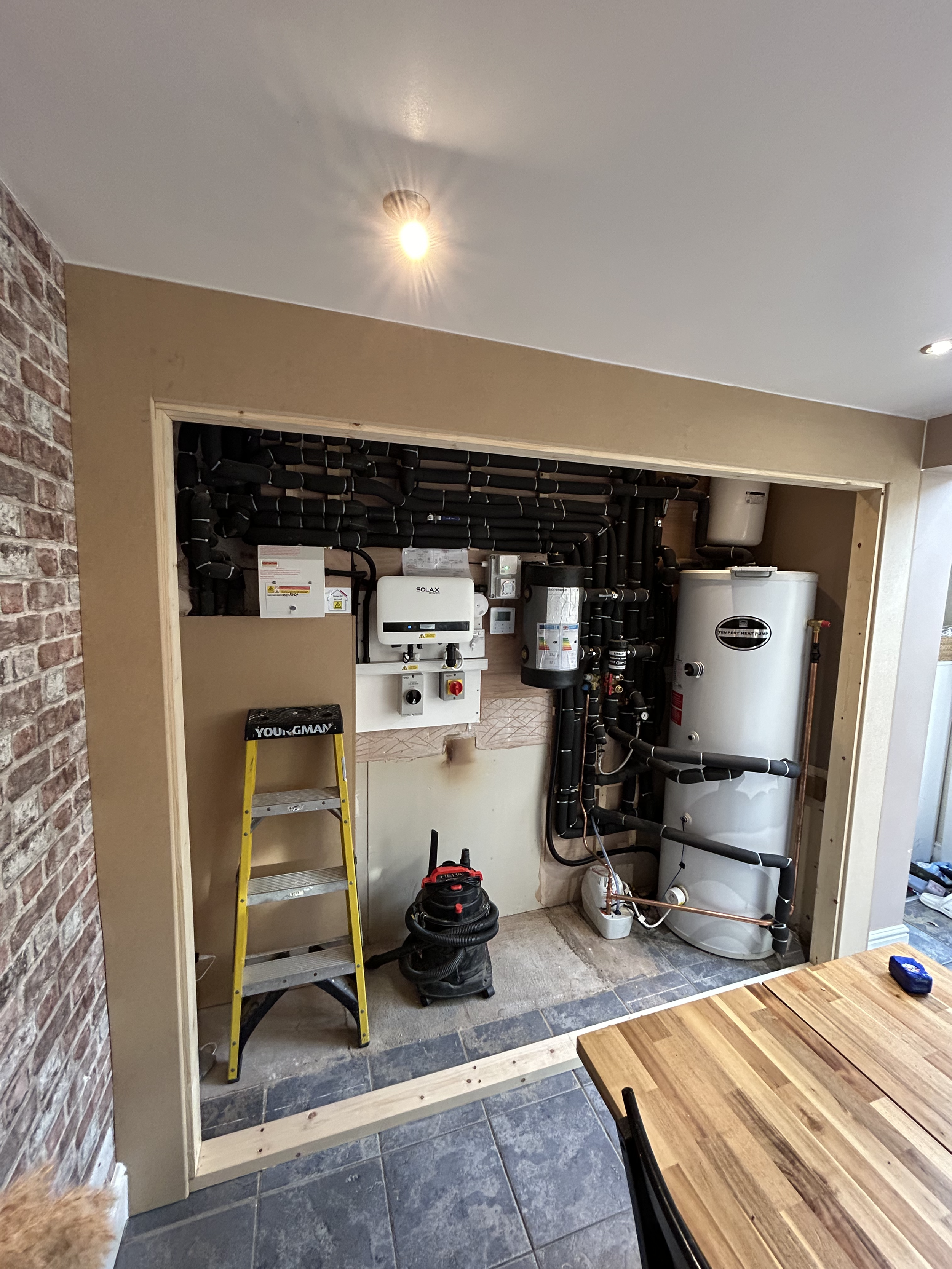 A partially constructed utility room with exposed plumbing and equipment, including water heater and heating system controls, a yellow ladder, and a black vacuum cleaner, adjacent to a brick wall and a wooden dining table.