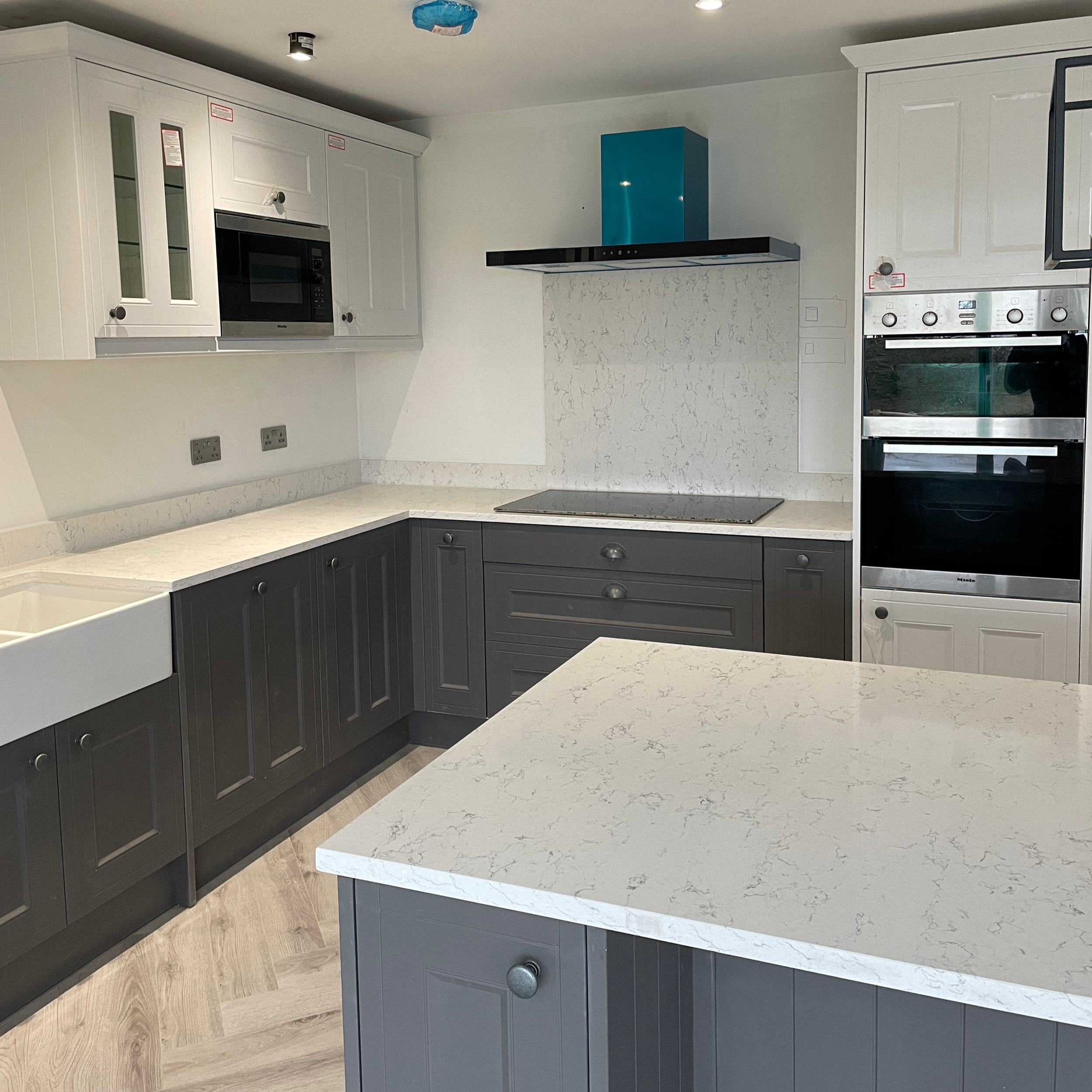 Modern kitchen with white upper cabinets, gray lower cabinets, a white marble countertop, a built-in microwave, stainless steel oven, and a blue range hood, with hardwood flooring.