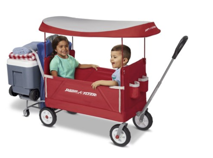Two children sitting in a red Radio Flyer wagon with a canopy, with a box of supplies attached to the back.