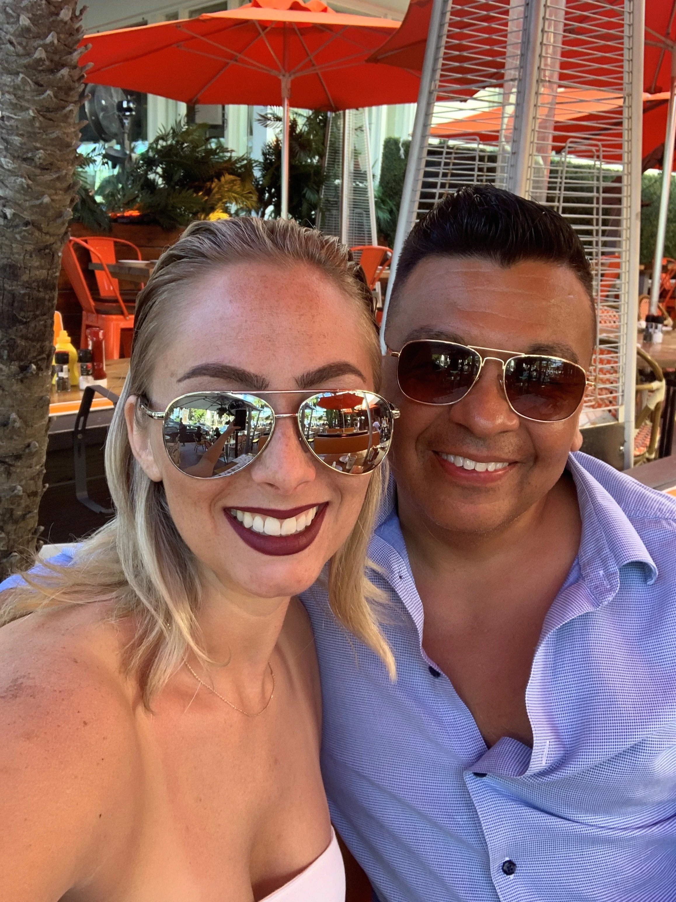 Close-up photo of a smiling woman and man, both wearing sunglasses, at an outdoor restaurant or cafe with orange umbrellas and greenery in the background.