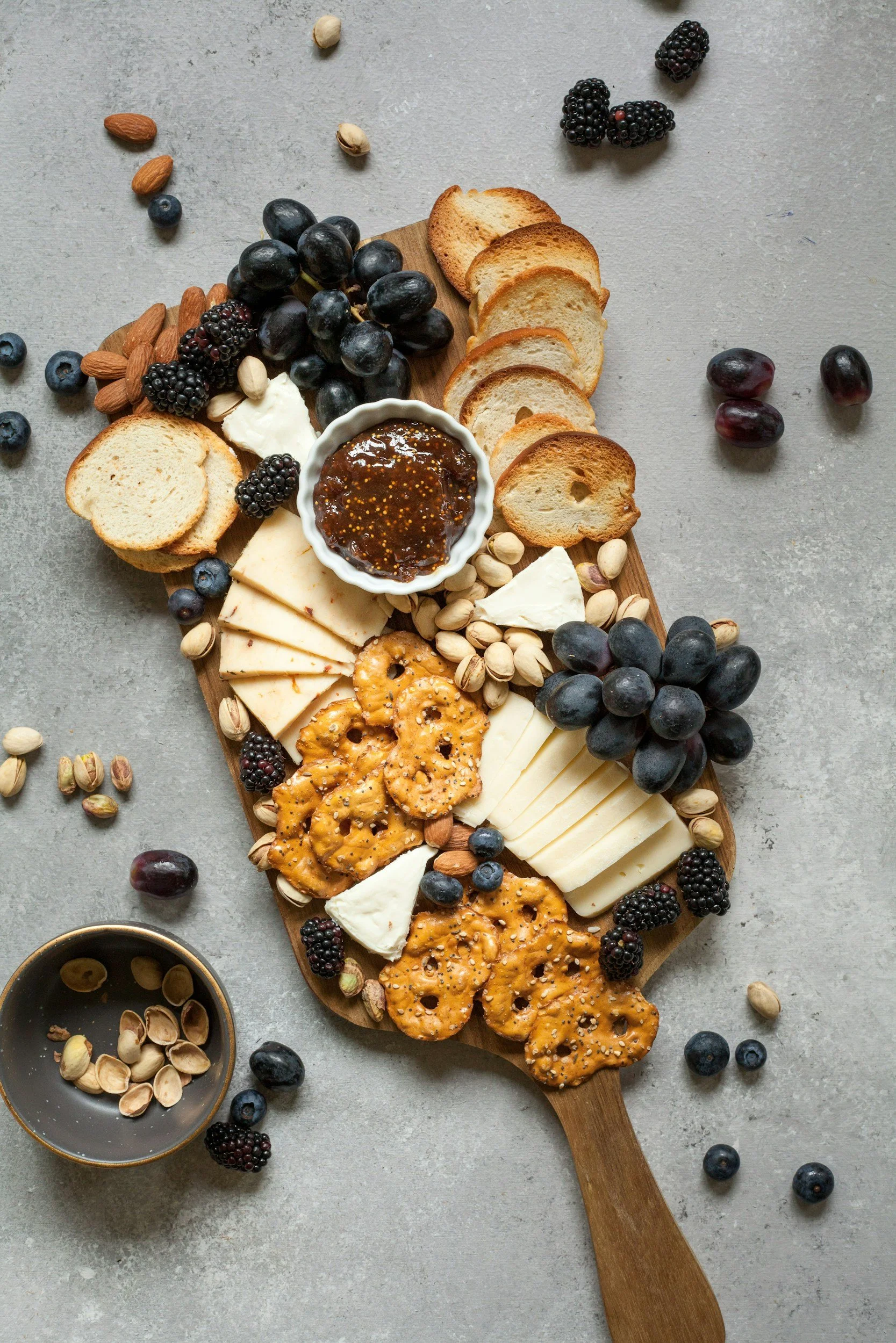 A wooden serving board with an assortment of cheeses, crackers, and fruits including blackberries, blueberries, black grapes, and pistachions. There is a small white bowl of jam or preserves. Scattered berries and nuts are also on the surface around the board.