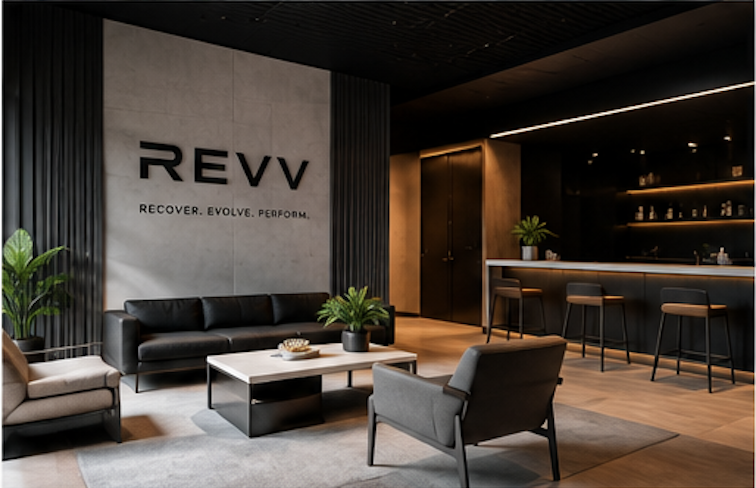 Modern lounge area with black sofa, armchairs, and coffee table, featuring the REVV logo and slogan, with plants and a bar counter in a stylish, dimly lit setting.