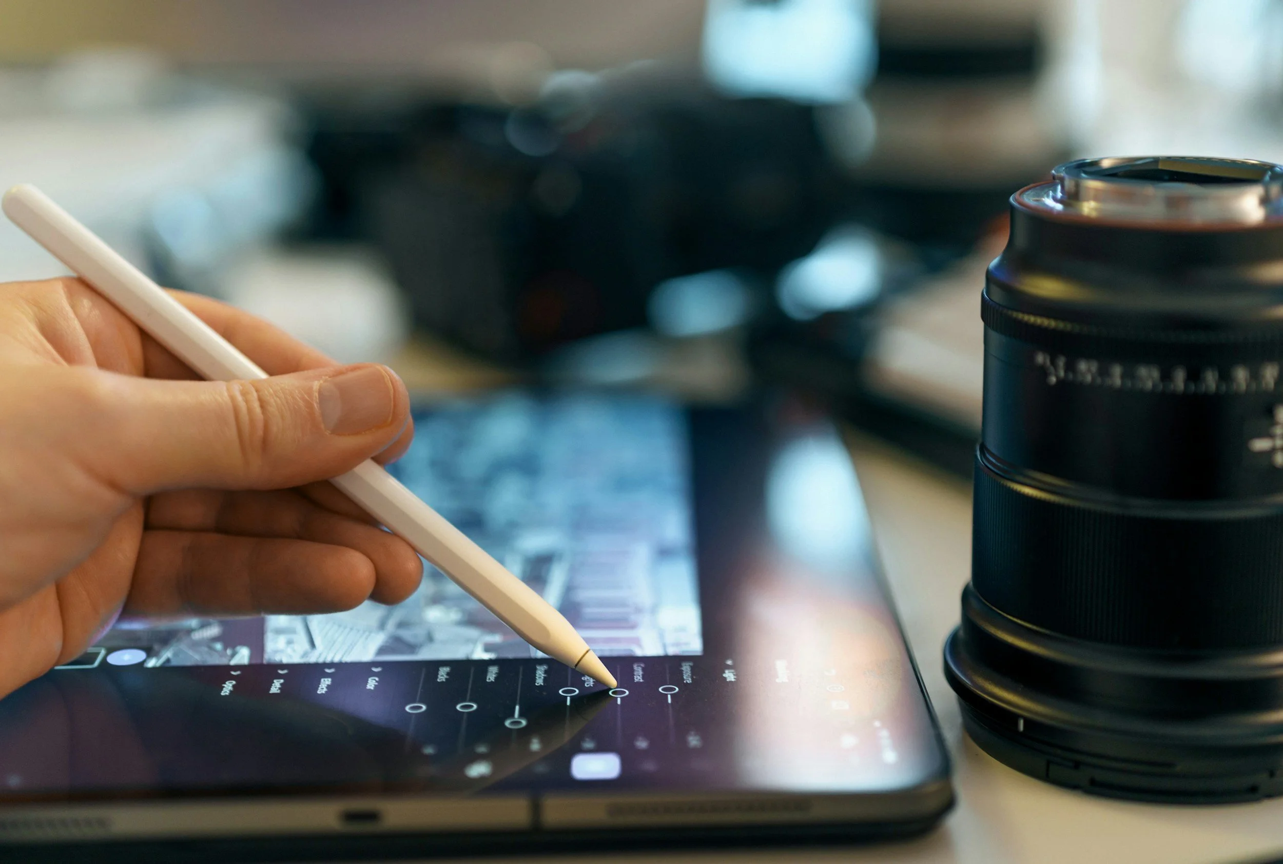Person holding a stylus on a digital tablet with photo editing or design app open, surrounded by a camera lens and an empty drink can.