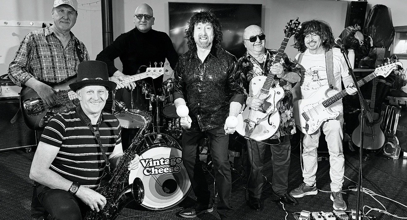A six-member band named Vintage Cheese performing indoors with musical instruments, including drums, electric guitars, and a saxophone, in a room with a television and various equipment.