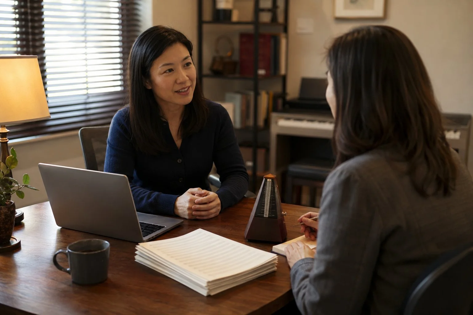 A specialty tutoring studio owner in a navy cardigan reviewing financials with a community lender across a desk, a metronome and a stack of blank sheet music beside the laptop, a keyboard visible behind