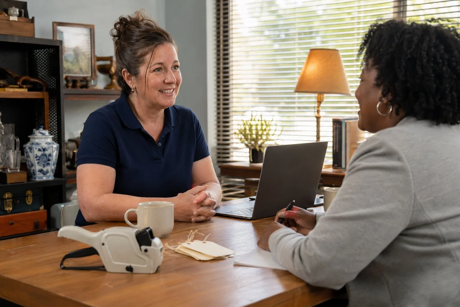 An estate sale professional in a navy polo reviewing financials with a community lender across a desk, a price-tag gun and inventory tags on the table