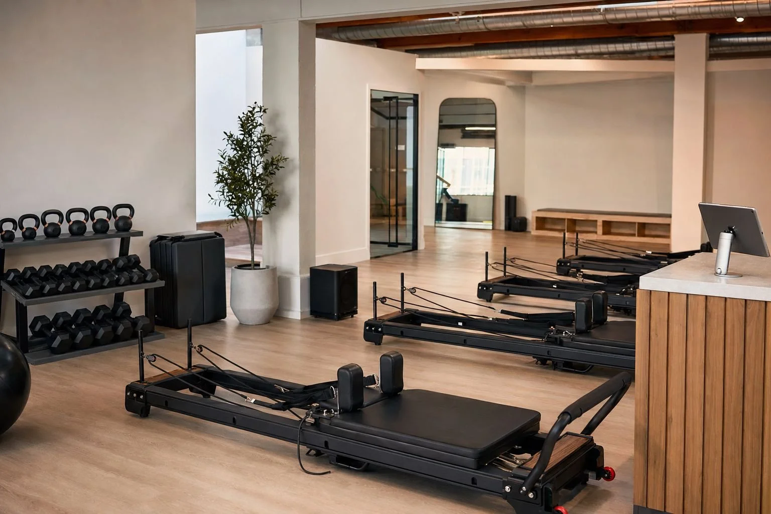 Boutique studio startup equipment in an open studio — reformers, racked kettlebells and dumbbells, stacked mats, a small speaker, and a front-desk tablet stand