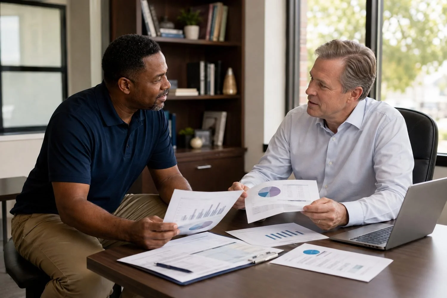 The same senior move specialist, in his navy polo and khakis, sitting at a table with a community lender in a light shirt, the two of them going through printed financial statements and a referral-source schedule together