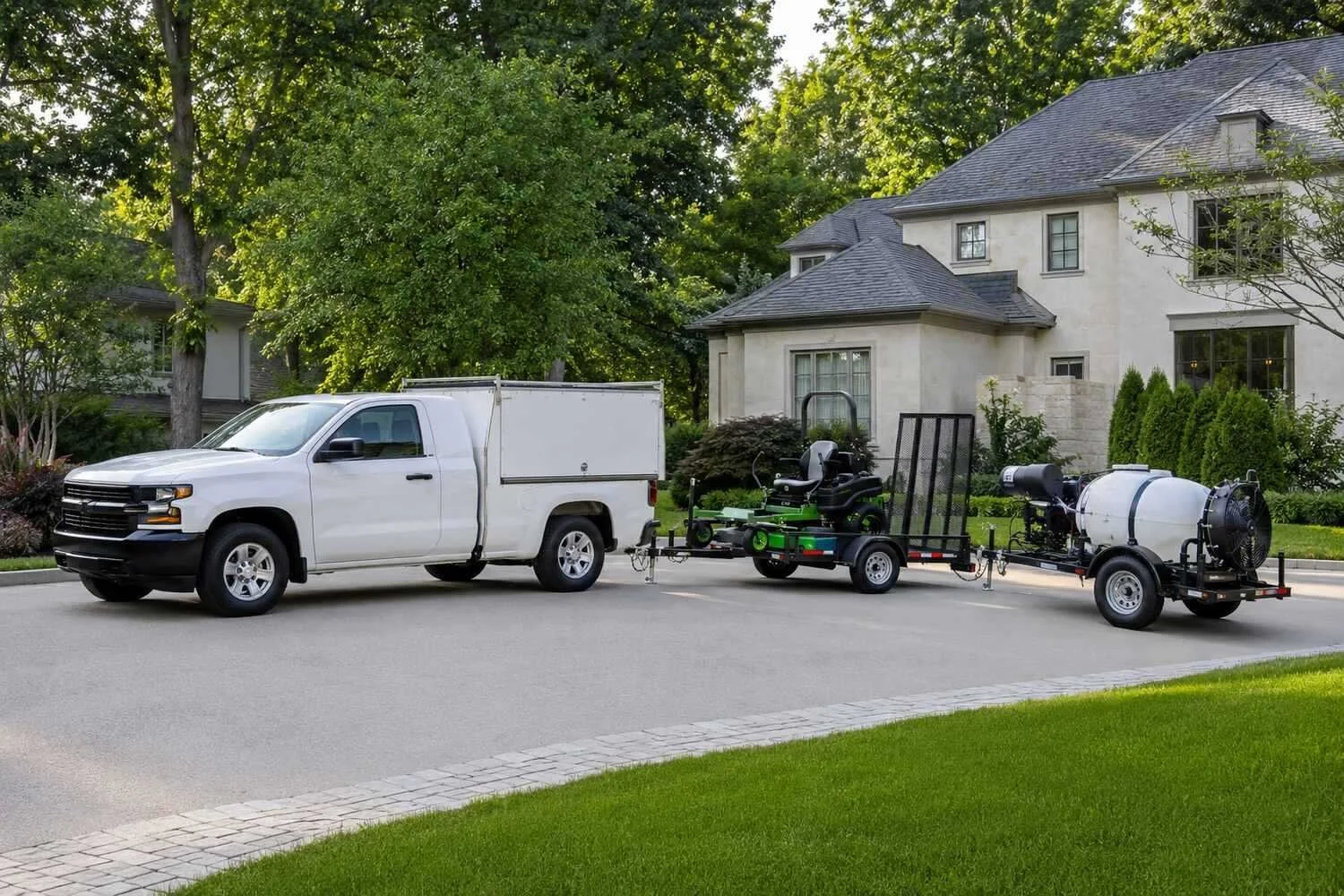 A white service pickup with a covered utility bed hitched to a trailer carrying a commercial zero-turn mower and a towed mosquito-control mister, parked together on a driveway — three route services running off one vehicle