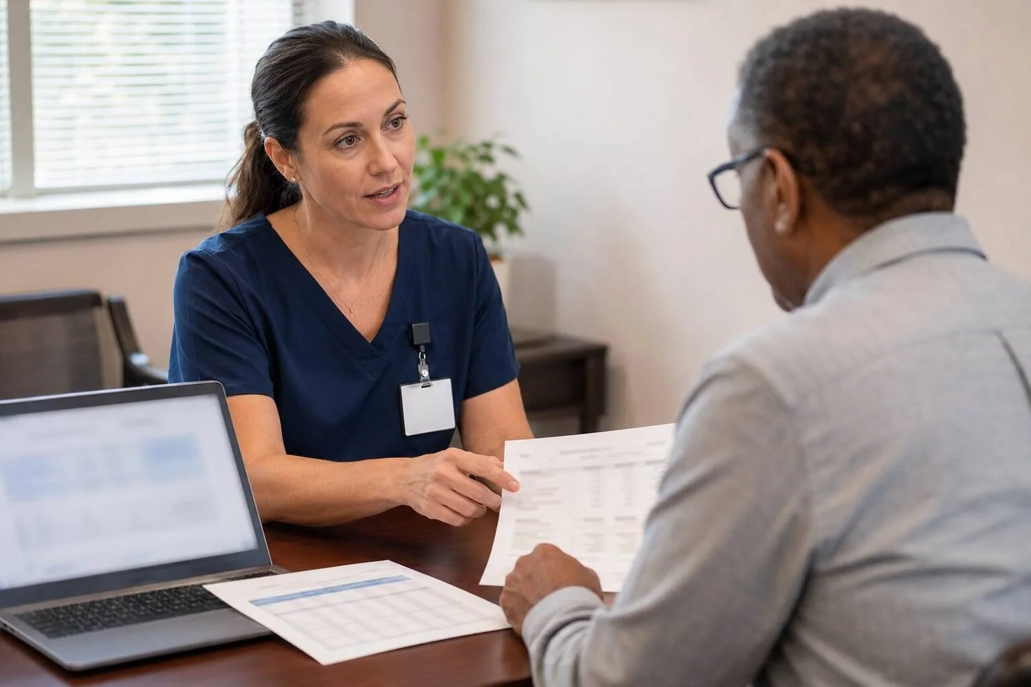 A senior care agency owner in a navy scrub top reviewing printed financial statements with a community lender across a desk, a staffing schedule and laptop between them