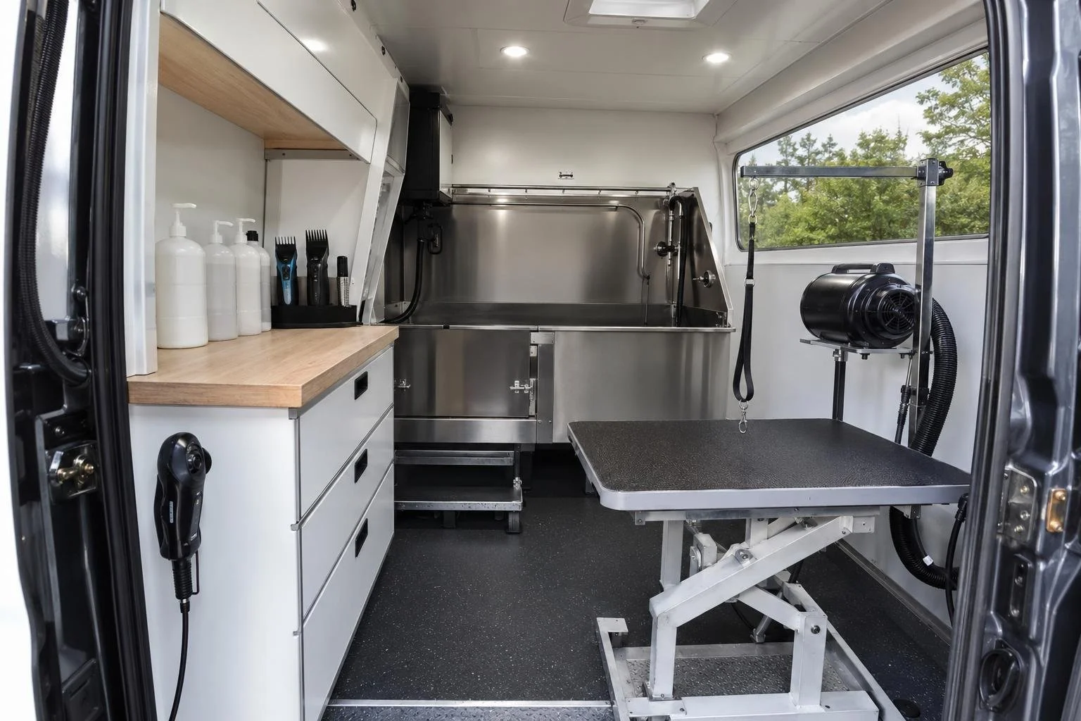 The interior of a grooming van conversion — a stainless hydraulic tub, an adjustable grooming table, a high-velocity dryer, clippers, and shampoo bottles on a fitted counter
