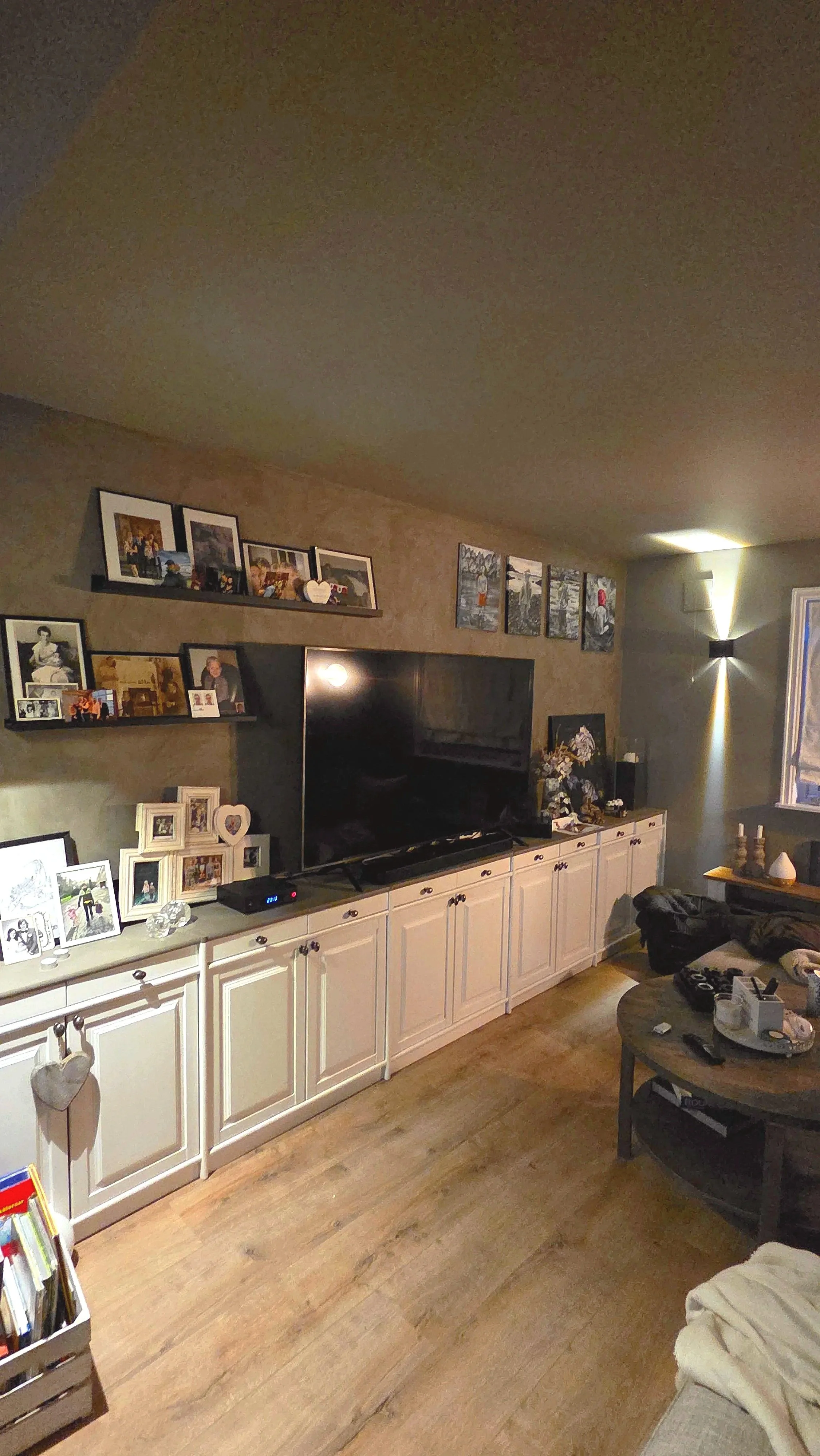Stue med hvite skap og sort TV, dekorert med bilder og dørmatte. Hylle med bilder og pynt over TV, veggkunst og lys. To sofaplasser med klær og ting, et rundt bord med fjernkontroller og andre småting.