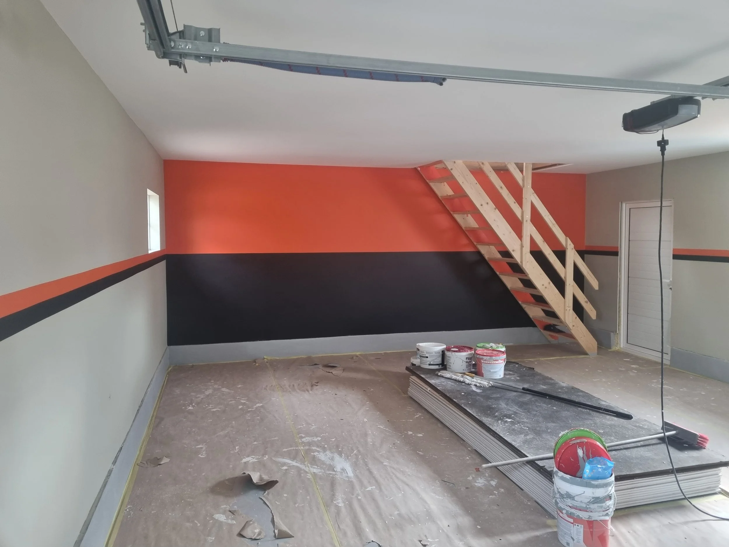 På et rom under oppussing er det maling, malerspann, og byggematerialer. Trapper fører opp til et loft eller andre rom, og det er et ferdig malt vegg med fargene mørk grå, oransje, og hvit.