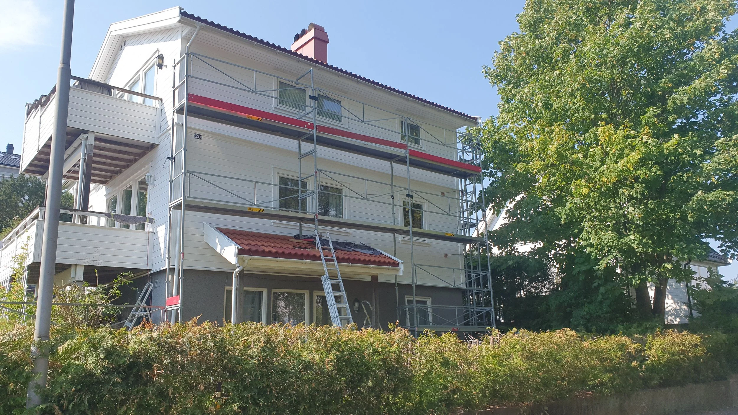 Et hvitt hus med tre etasjer og balkonger, omringet av grønt løvtrær, med byggestillas foran.