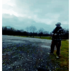 A person standing outdoors on a gravelly surface near a grassy area under a cloudy sky, wearing a backpack and a hat.
