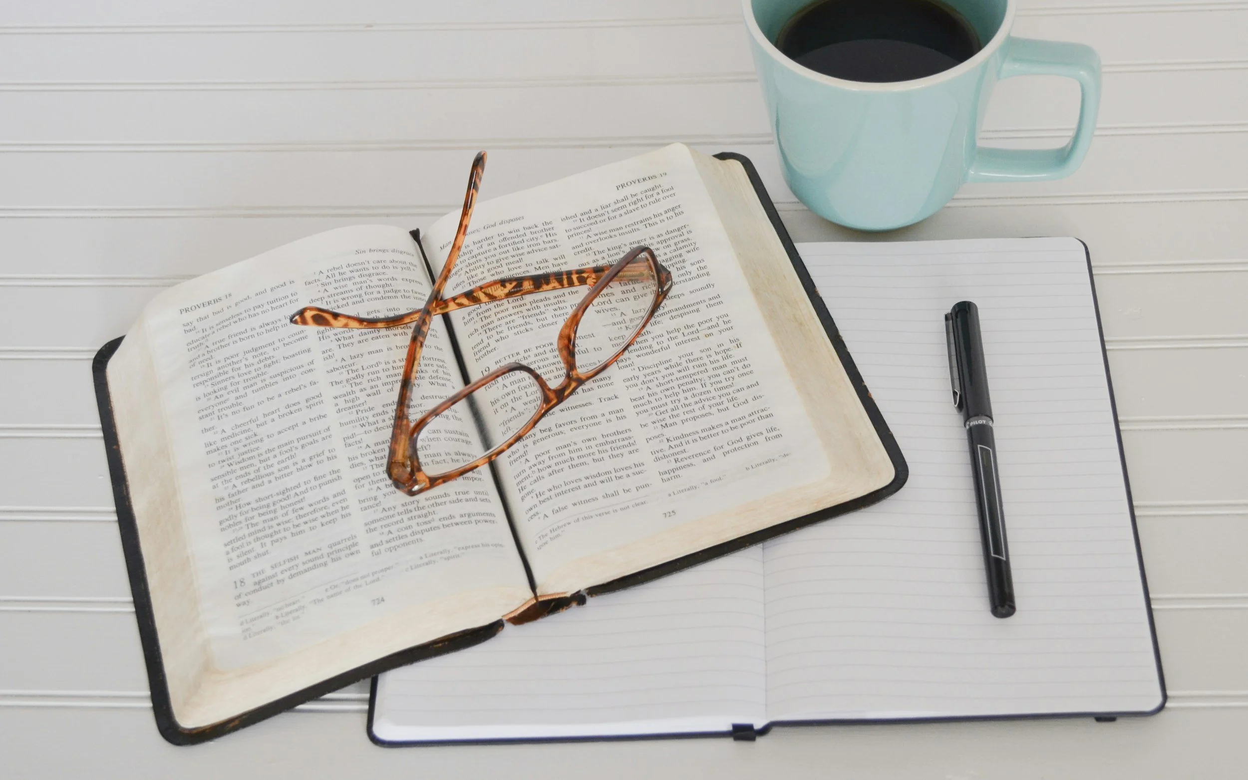 Open Bible with tortoiseshell glasses resting on it, a black pen, notepad, and a light blue coffee mug filled with dark coffee on a white striped surface.