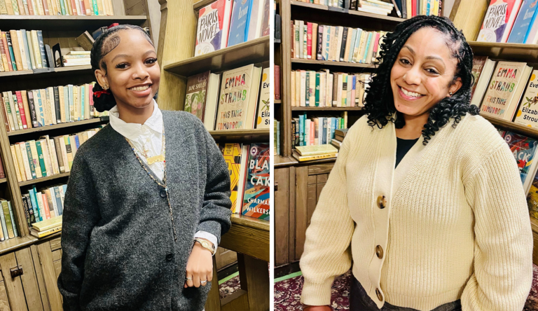 Two women smiling in front of bookshelves in a bookstore or library, one wearing a gray cardigan and the other in a cream sweater.