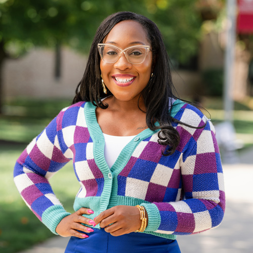 Woman wearing glasses and a colorful checkered cardigan smiling outside in a park.