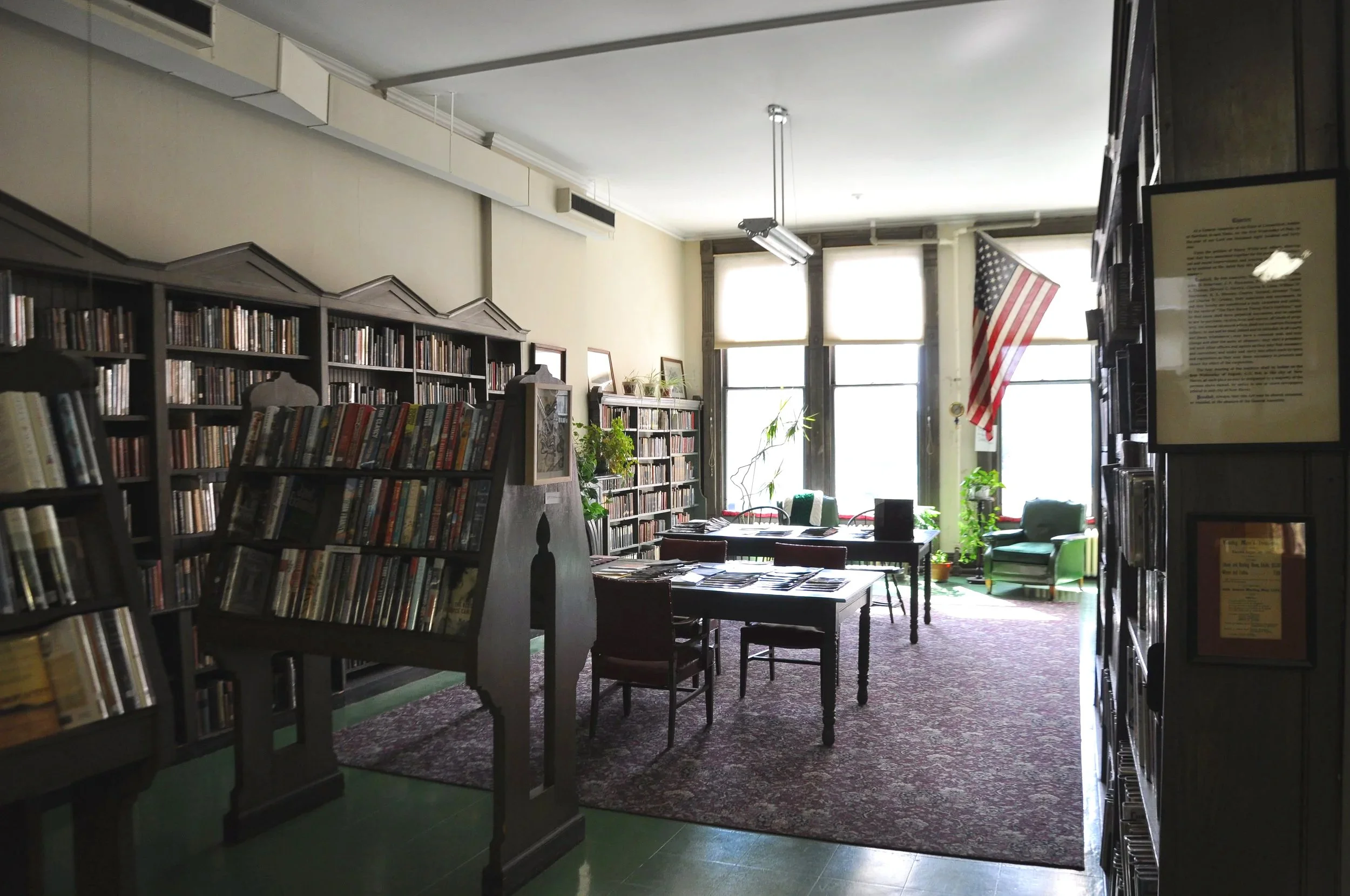 Interior of a library or bookstore with shelves filled with books, tables, chairs, and large windows providing natural light, American flag hanging near the window.