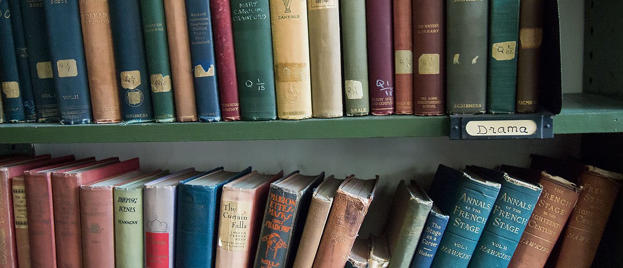 Books organized on a library shelf, labeled with a handwritten sign that says 'Drama' and other books on different topics.