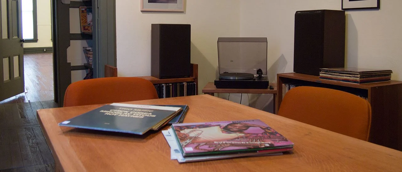 A wooden dining table with several vinyl records and magazines, orange chairs around it, and a record player with speakers in the background, in a cozy room.