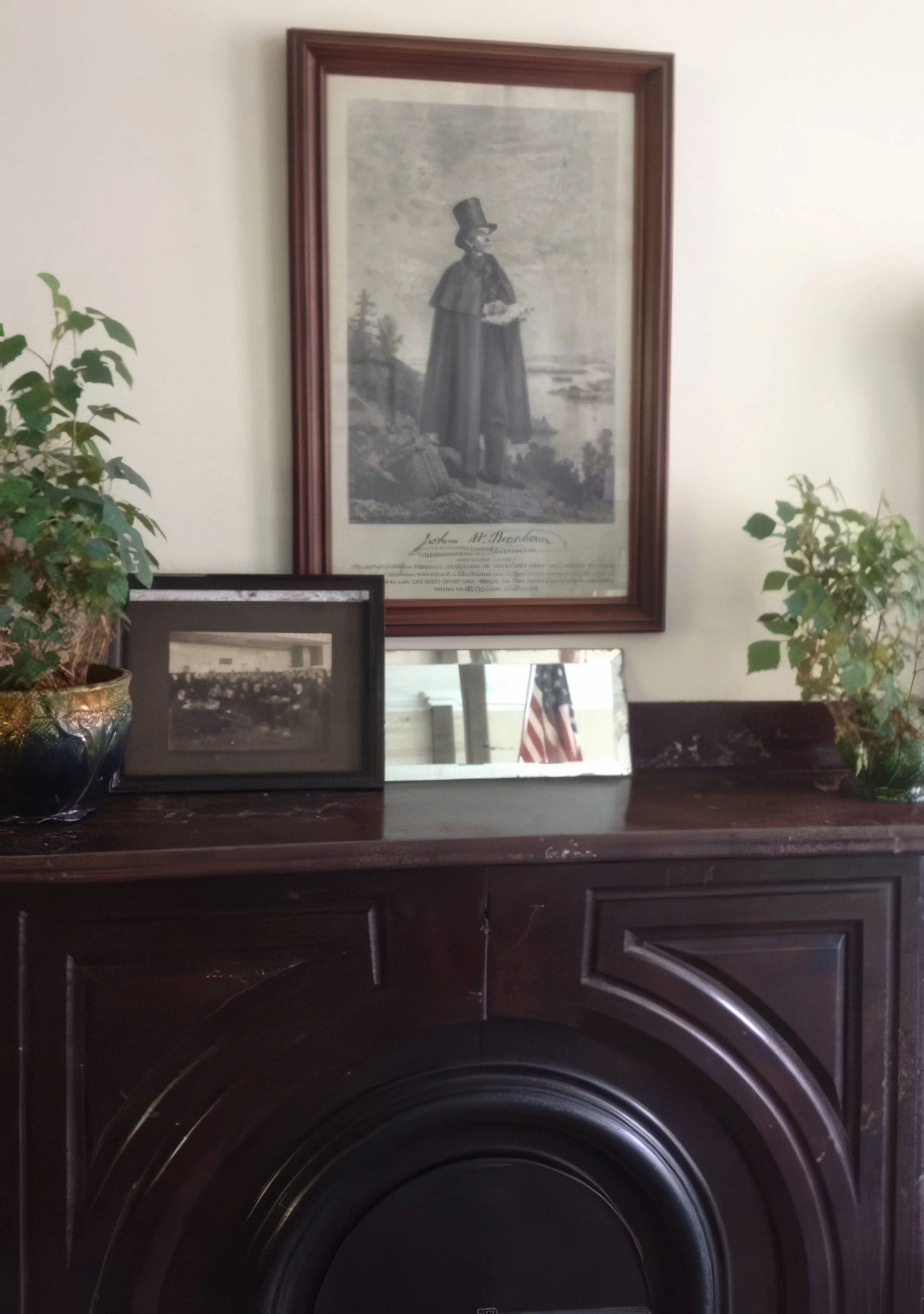 Framed black and white print of a woman in Victorian attire standing outdoors with river and trees in the background, on a wall above a dark wooden surface, with two potted plants and two framed photographs in front.