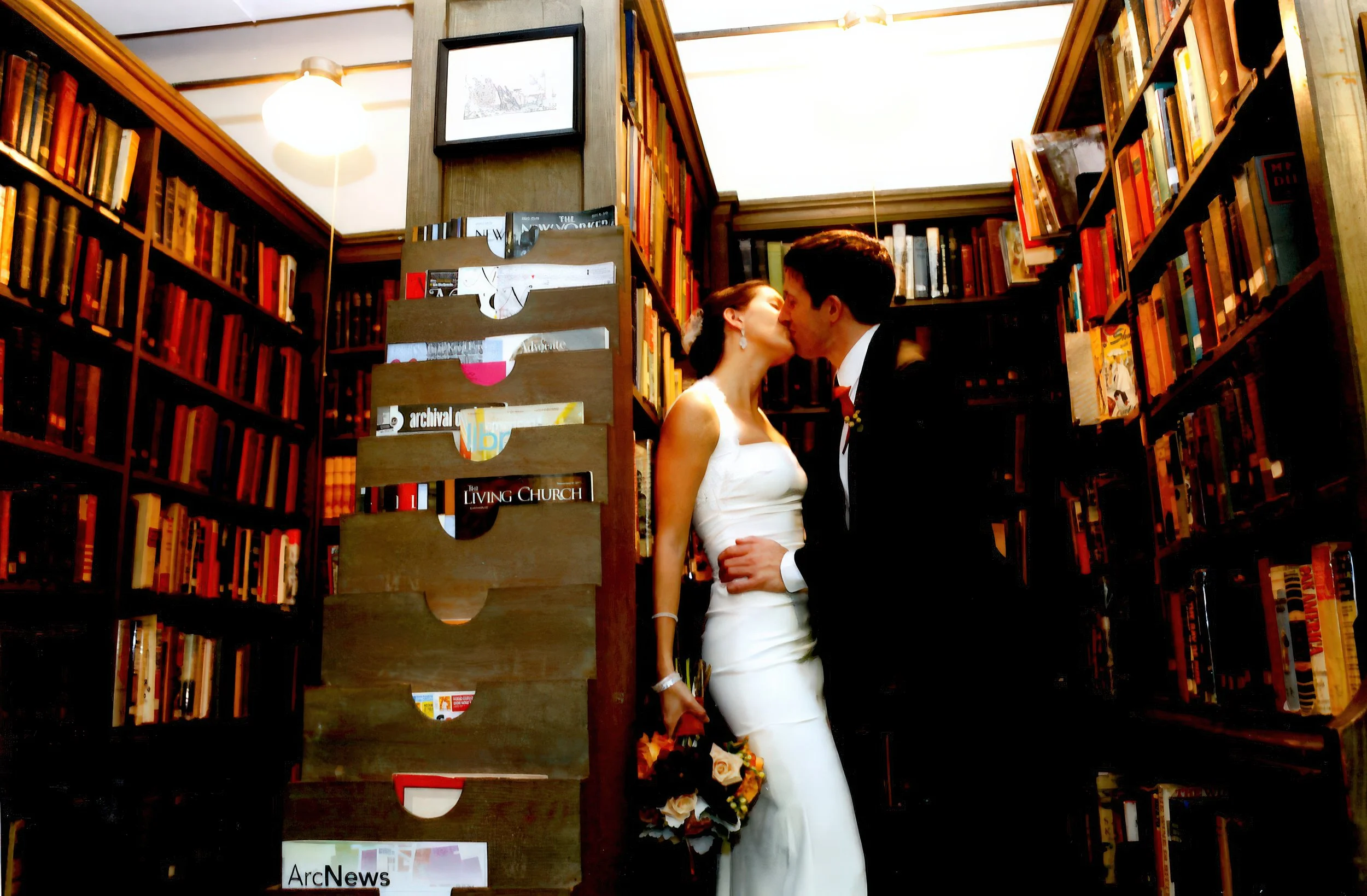 A bride and groom share a kiss in a library surrounded by tall bookshelves filled with books.