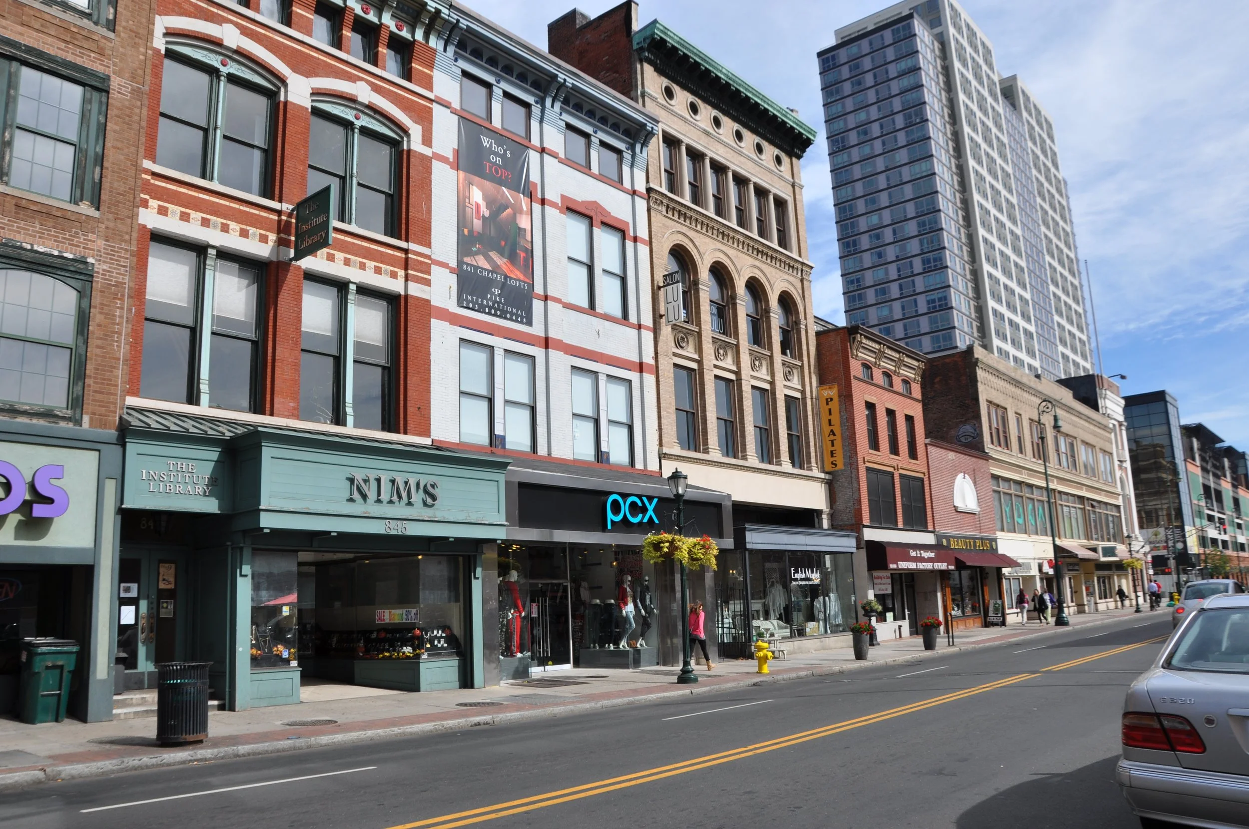 A city street with multiple multi-story buildings, including a library named The Institute Library, storefronts, and retail shops. The street has parked and moving cars, streetlights, and street signs. The sky is partly cloudy.
