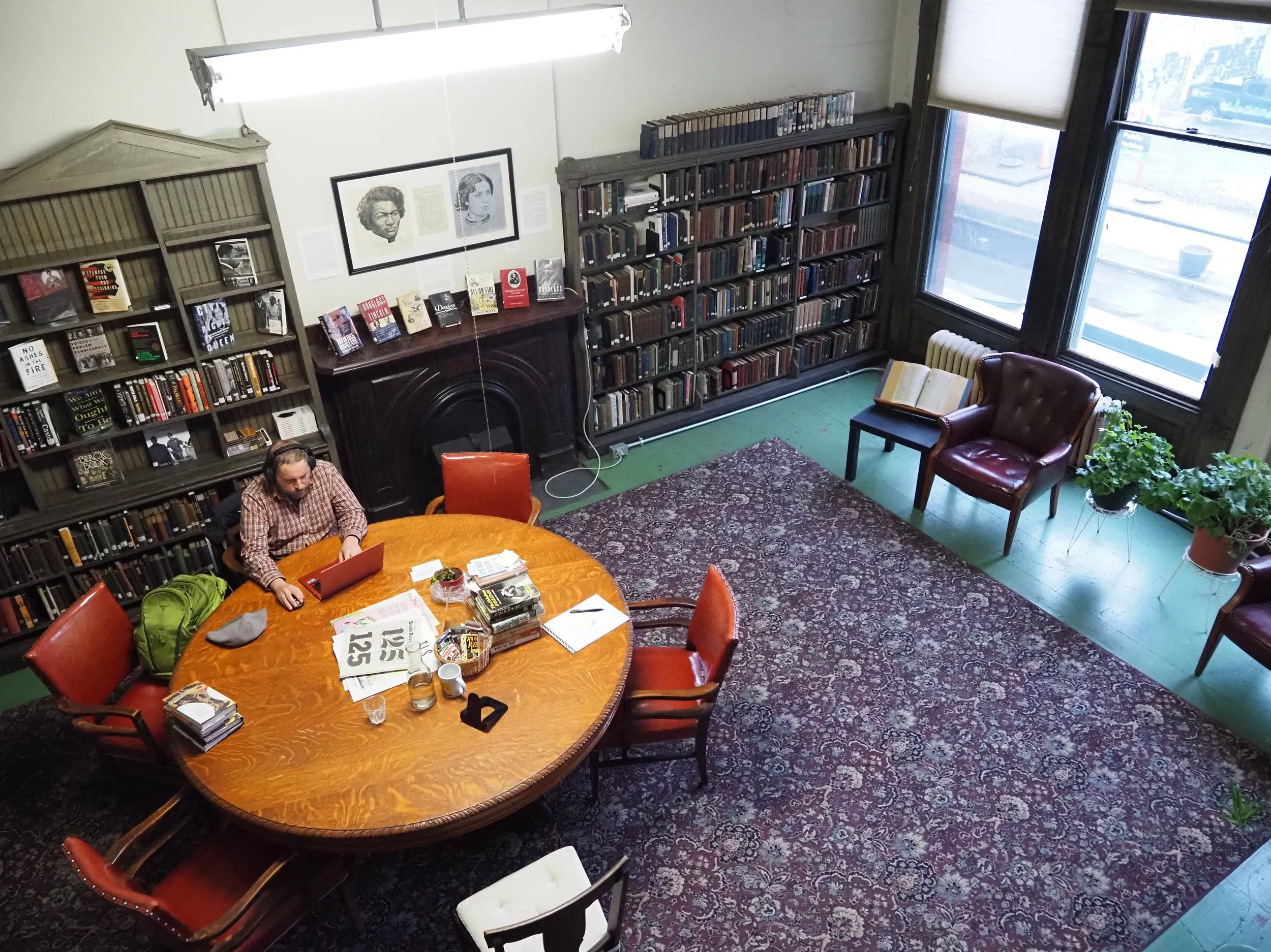 A cozy bookstore or library room with tall shelves filled with books, a large round wooden table with red chairs, a person working on a laptop, large windows with blinds letting in natural light, a patterned rug, a potted plant, and framed portraits 