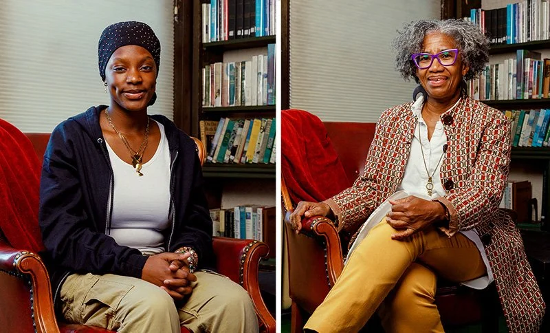 Two women sitting in front of bookshelves in a library, each on a red armchair. The woman on the left is younger, wearing a black hoodie, khaki pants, a black beanie with white dots, and jewelry. The woman on the right is older, with curly gray hair, wearing glasses, a patterned blazer, white shirt, and yellow pants.