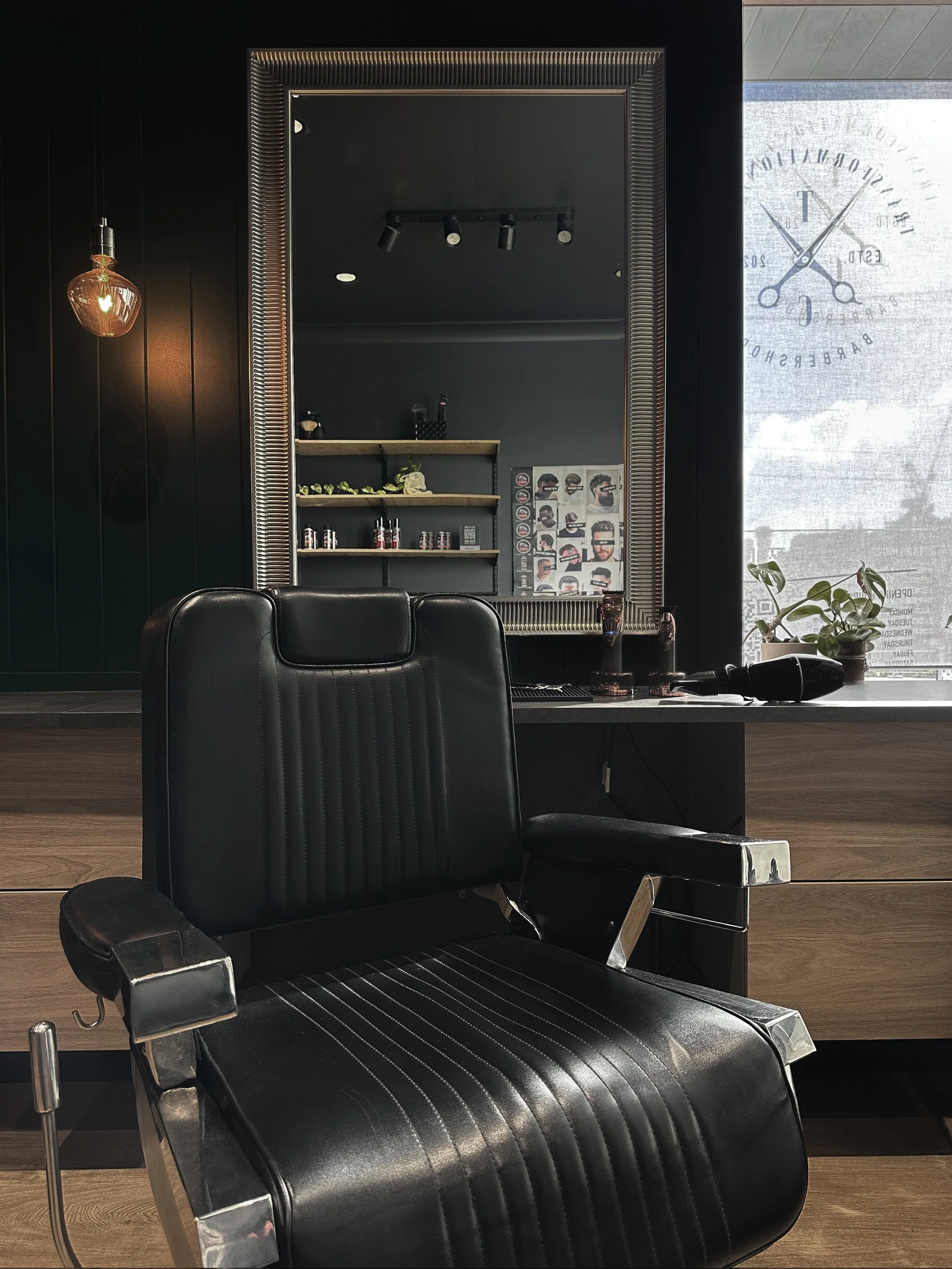 A black barber chair with a high backrest and chrome armrests in a modern barber shop. Behind, a large mirror with a decorative frame, a wooden shelf displaying small bottles, plants, and a wall collage of photos. To the left, a hanging light bulb provides warm lighting, and a window on the right lets in daylight with a large clock and a plant on the windowsill.