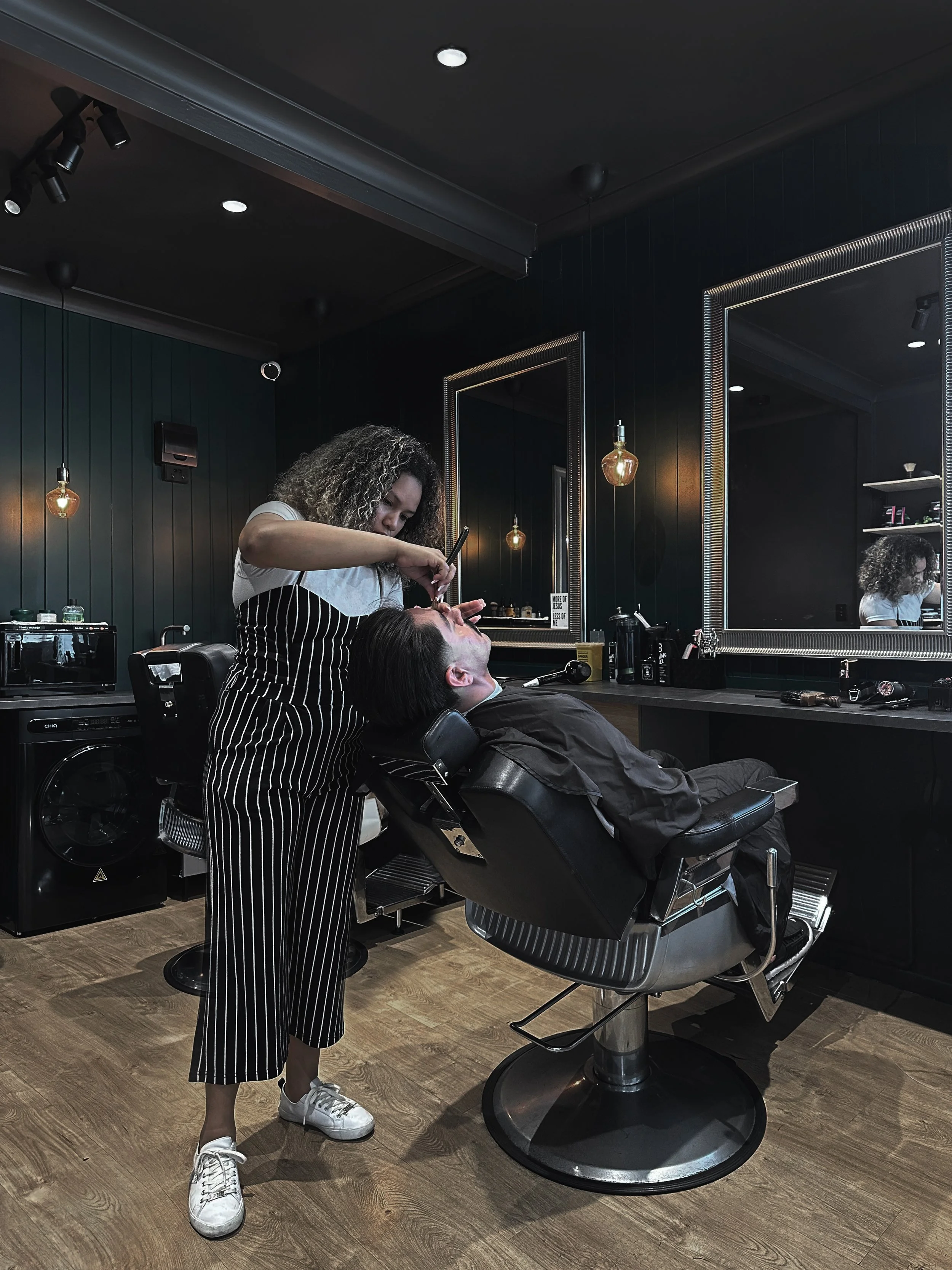 A woman is giving a man a haircut at a modern barbershop or salon. The man is reclining in a black barber chair while the woman stands next to him, focused on her work. There are large mirrors on the dark-colored wall, decorative lightbulbs, and various barber tools on the counter.