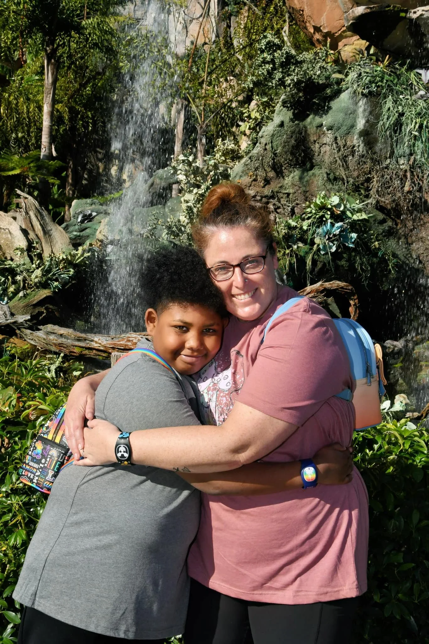 A woman and a young boy hugging each other outdoors near a waterfall with lush green plants and rocks in the background.