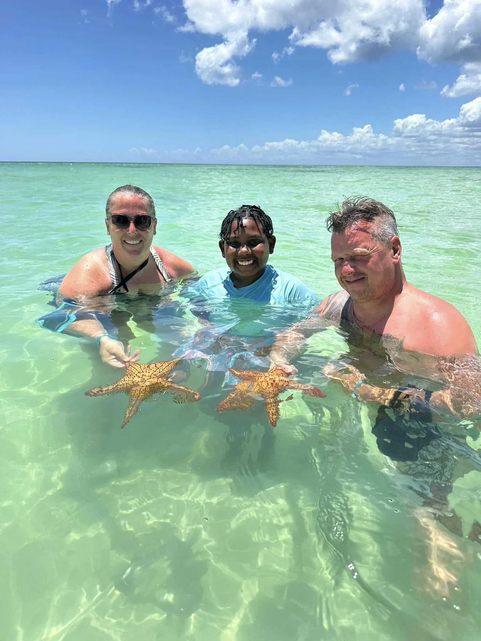 Three people smiling in clear, shallow ocean water with starfish visible below the surface. Blue sky with clouds above.