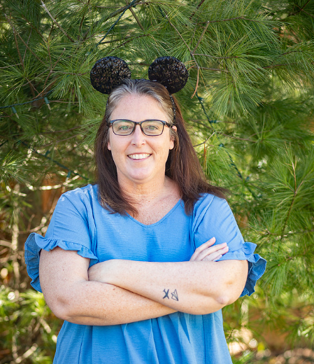 Woman wearing glasses and a blue shirt with ruffled sleeves, standing outdoors in front of green pine tree, with Mickey Mouse ears headband.