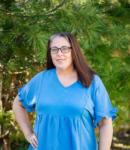 Woman with glasses and brown hair wearing a blue dress standing outdoors in front of green trees.